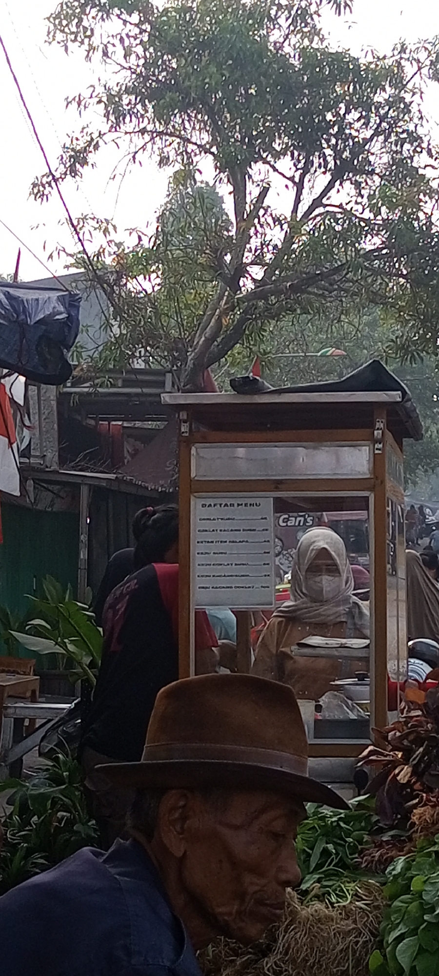 In Setu, Indonesia, a man wearing a brown fedora and a dark blue shirt is seen in the foreground. He is looking down, with his face partially obscured by shadows. Behind him, a food stall with a "DAFTAR MENU" sign is visible. A woman wearing a light brown hijab and a face mask is standing behind the counter, serving customers. The stall has a display of food items and is located outdoors, with trees and other stalls in the background. The scene appears to be a busy market or street food area.