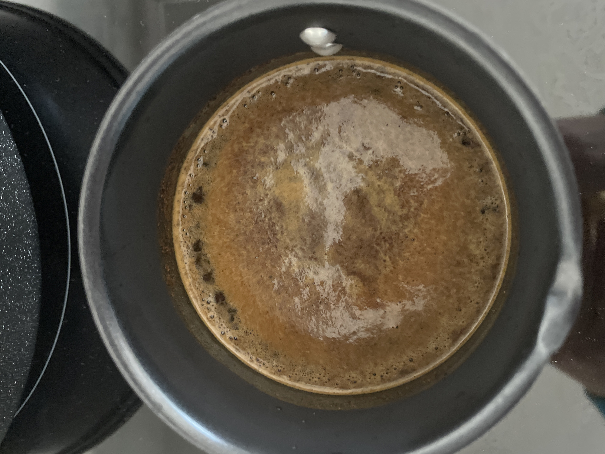A top-down view shows a Moka pot filled with freshly brewed coffee. The coffee has a frothy, light brown surface with darker brown edges. The Moka pot itself is grey metal, and it sits on a black stovetop. There is a small amount of white residue or substance on the inside rim of the pot. It is difficult to determine the exact location.