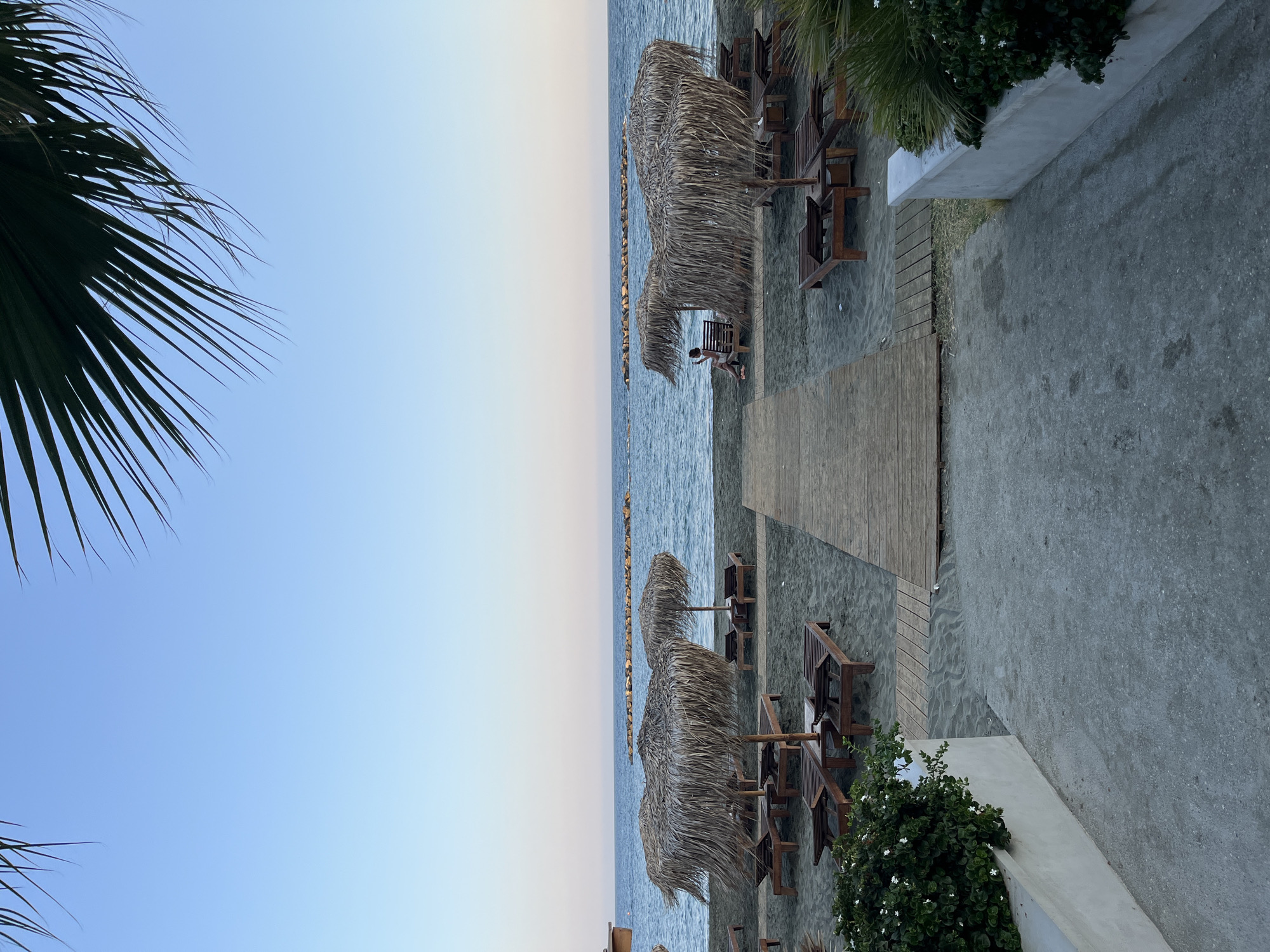 The image depicts a serene beach scene in Agios Tychona Community, Cyprus. In the foreground, a grey concrete path leads down towards the beach. To the left, a white planter box holds lush green bushes with small white flowers. To the right, another white planter box contains similar foliage and a palm frond.
The beach itself is sandy and appears dark. Several wooden lounge chairs with straw umbrellas are set up, with some unoccupied and others with lounge chairs empty. A wooden boardwalk extends across the sand.
The sea is visible in the mid-ground, with a calm surface and a clear horizon. A line of rocks forms a breakwater in the distance. A lone figure, a woman wearing a light-colored dress, is seated on a lounge chair under a straw umbrella on the right side of the beach, appearing to be looking at her phone.
The sky above is a clear, pale blue, with the dark fronds of a palm tree visible in the upper corners of the frame, suggesting the time of day is late afternoon or early evening. The overall atmosphere is peaceful and tranquil, characteristic of a quiet beach resort.