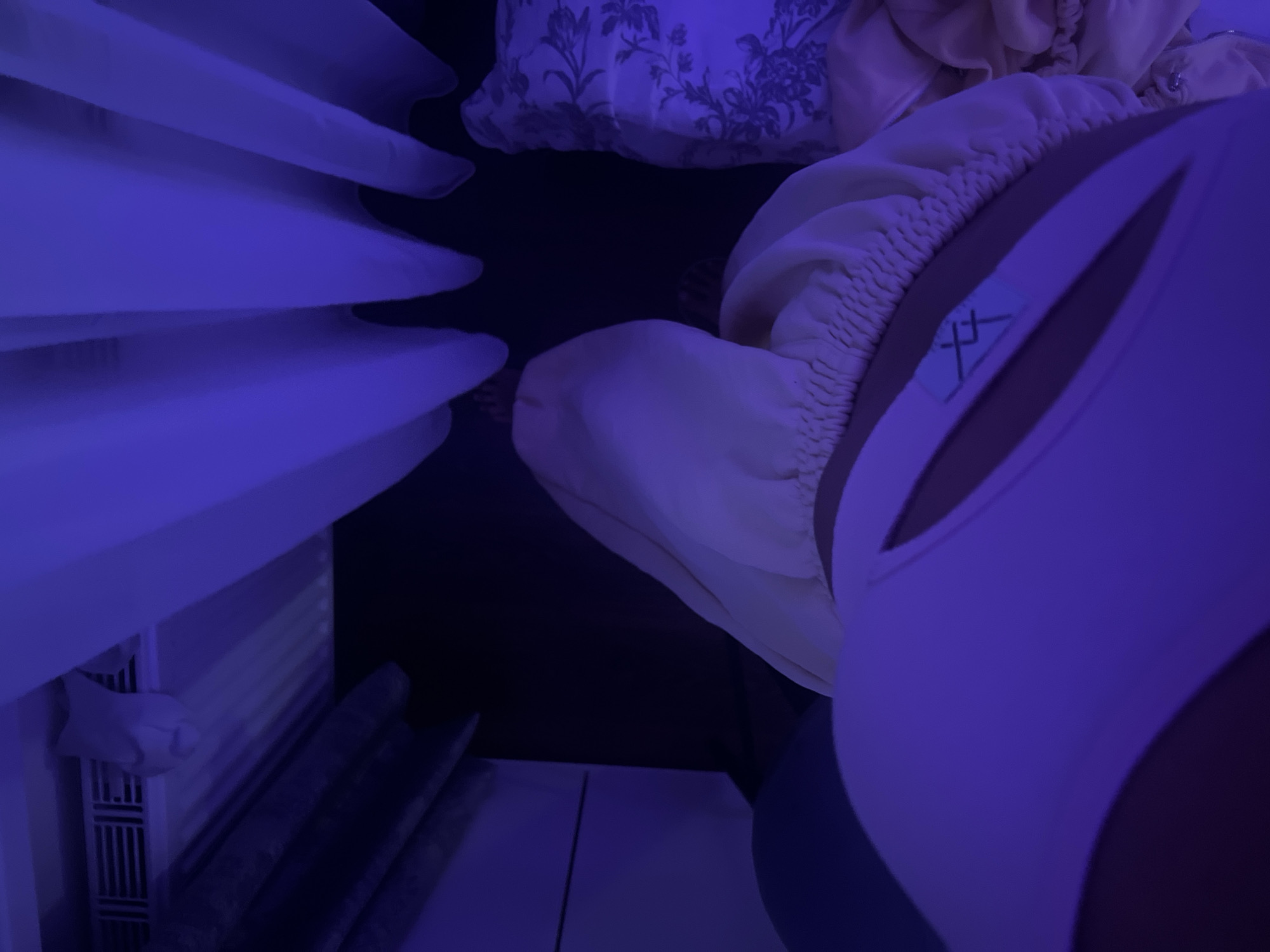 The image shows a close-up, low-angle shot of a person wearing a white garment with a blue design on a tag. The garment appears to be part of a swimsuit or activewear. The person is lying down, and a cream-colored bonnet or cap is visible on their head. The background includes draped white fabric, possibly curtains or a canopy, and a pillow with a floral pattern. The lighting is predominantly purple, creating a moody and somewhat abstract atmosphere. The scene is indoors, and no identifiable elements suggest a specific location in Saint Petersburg, Russia.