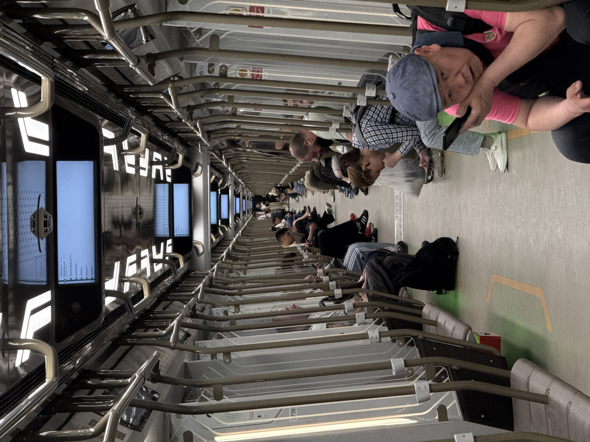 The image shows the interior of a sleeper car on the Aeroexpress train from Moscow to Sheremetyevo Airport in Moscow, Russia. Passengers are reclining on individual bunk beds, many of whom are using their phones or resting. The train car has multiple berths arranged in rows along each side. Information screens are visible at the end of the car. The carriage is well-lit, with metal rails and fittings. The atmosphere is one of relaxed travel, with passengers appearing comfortable and at ease during their journey.