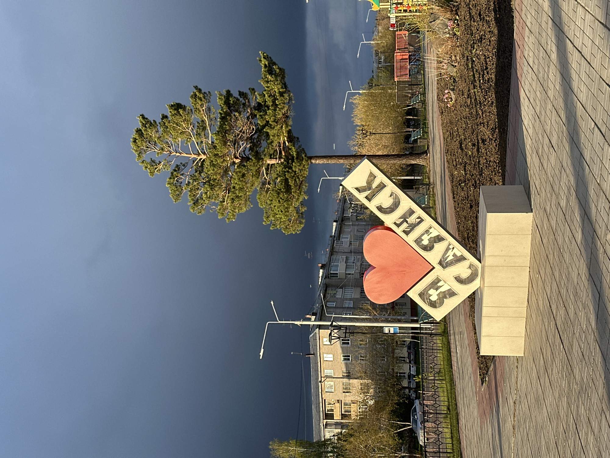 The image shows a view in Sayansk, Russia, with a prominent "I <3 SAYANSK" sign in the foreground. The sign features a red heart shape and Russian text. A tree stands adjacent to the sign, and behind it are multi-story apartment buildings and streetlights. The sky in the background is a mix of dark, stormy clouds and brighter areas. The foreground consists of a paved walkway. A small playground area can be seen further in the background to the right.