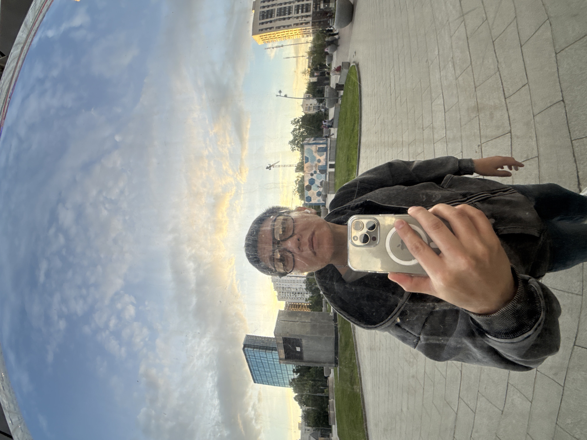 The image is a selfie reflection captured in a large, curved mirrored surface, appearing rotated approximately 90 degrees clockwise. The person taking the photo is visible from the chest up, wearing clear-rimmed glasses and a dark, possibly distressed, zip-up hoodie or jacket. They are holding a light-colored smartphone with a transparent case, which clearly shows a MagSafe ring and a triple camera array. A faint Apple logo is discernible on the phone case. The person’s expression is neutral as they gaze towards the camera.
The background, reflected and slightly distorted by the curved surface, presents an urban landscape consistent with Moscow, Russia. The sky, appearing on the left side of the image, features scattered white and grey clouds against a light blue backdrop, hinting at either late afternoon or early evening due to a soft, warm glow on some clouds. Below the sky, a mix of modern and older-style buildings can be seen, including a tall residential structure, a building with extensive blue-tinted glass, and another concrete-faced edifice. Two construction cranes are visible in the distance.
On the right side of the reflection, there is a paved area with light-colored rectangular tiles, a section of neatly manicured green grass, and several contemporary, block-like benches or seating arrangements. Streetlights and trees are also part of the urban setting, contributing to the impression of a public square or park area.