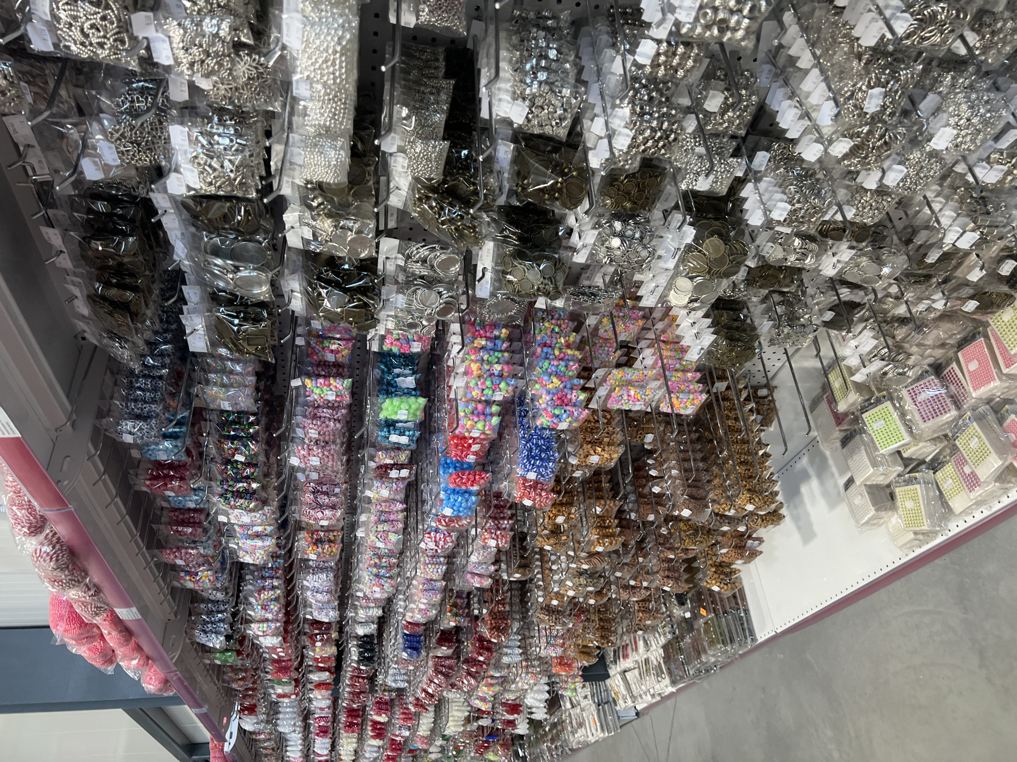 The image depicts a retail display, likely inside a store. It shows a wide selection of craft and jewelry-making supplies arranged on racks. The items are primarily small beads, charms, and findings packaged in clear plastic bags and hung from metal hooks or pegs. The items vary in color, material, and design. The location is Burgas, Bulgaria.