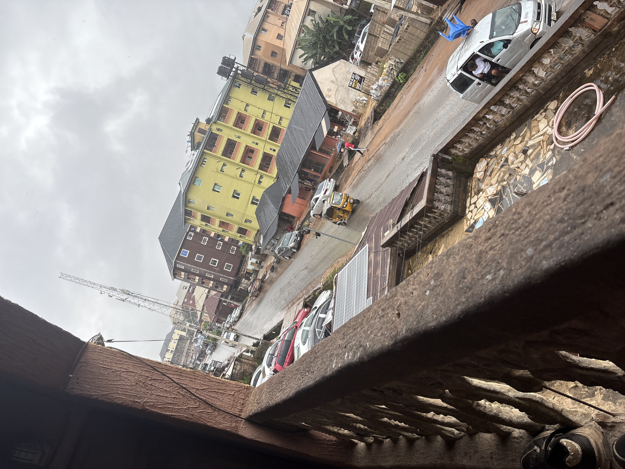 The image captures a street scene in Nkpor, Nigeria, on an overcast day, likely after rainfall, as indicated by the wet pavement and puddles. The perspective is from an elevated position, looking down a street lined with multi-story buildings of various colors, including a prominent yellow building with red balconies. A large cell tower is visible in the background.
In the foreground, a rusty balcony railing and part of a textured wall create a framing element for the view. Below the railing, a tiled patio area is visible, with a coiled pink hose lying on the wet surface.
The street itself is moderately busy with vehicular traffic. Several cars are parked along the roadside. A yellow tricycle (Keke Napep) is moving along the street, and a silver minivan with two individuals visible inside is also present. A blue plastic chair is perched on top of the minivan. A person in a red shirt and white pants is walking on the street.
The environment appears to be a commercial and residential area. Some buildings have storefronts at ground level. Construction materials, including blocks and bags of sand, are piled near one of the buildings. The overall impression is of an active urban environment, possibly experiencing challenging weather conditions.