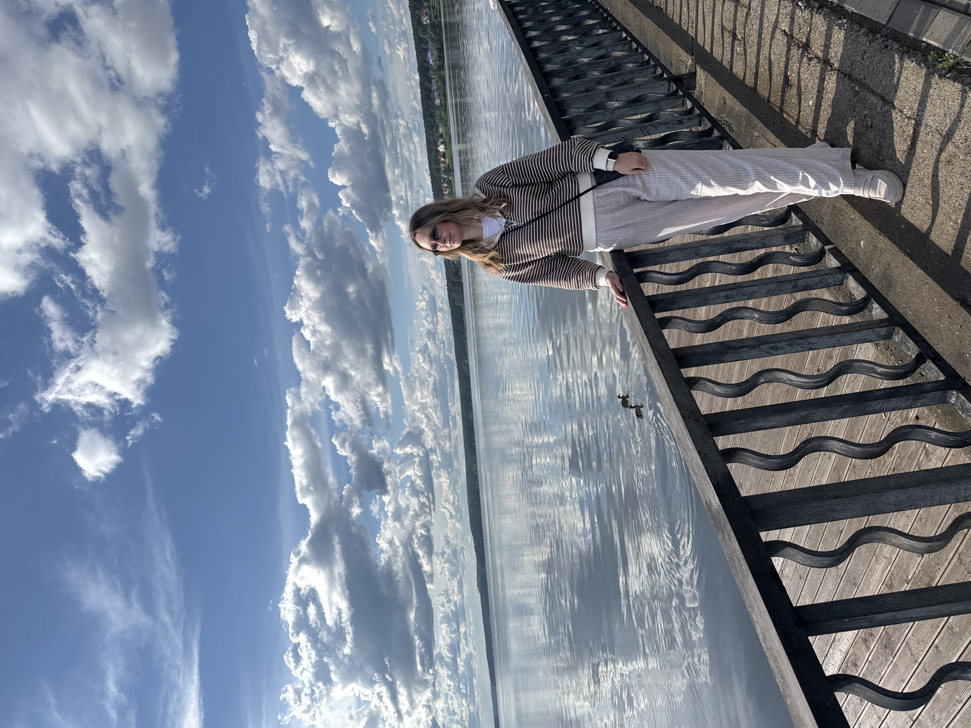 A full shot captures a person standing on a structure that appears to be a staircase leading into a body of water. The individual, dressed in striped pants and a matching sweater, is positioned at an angle. The body of water reflects the sky, which is filled with puffy clouds. The photo was taken in Votkinsk, Russia.