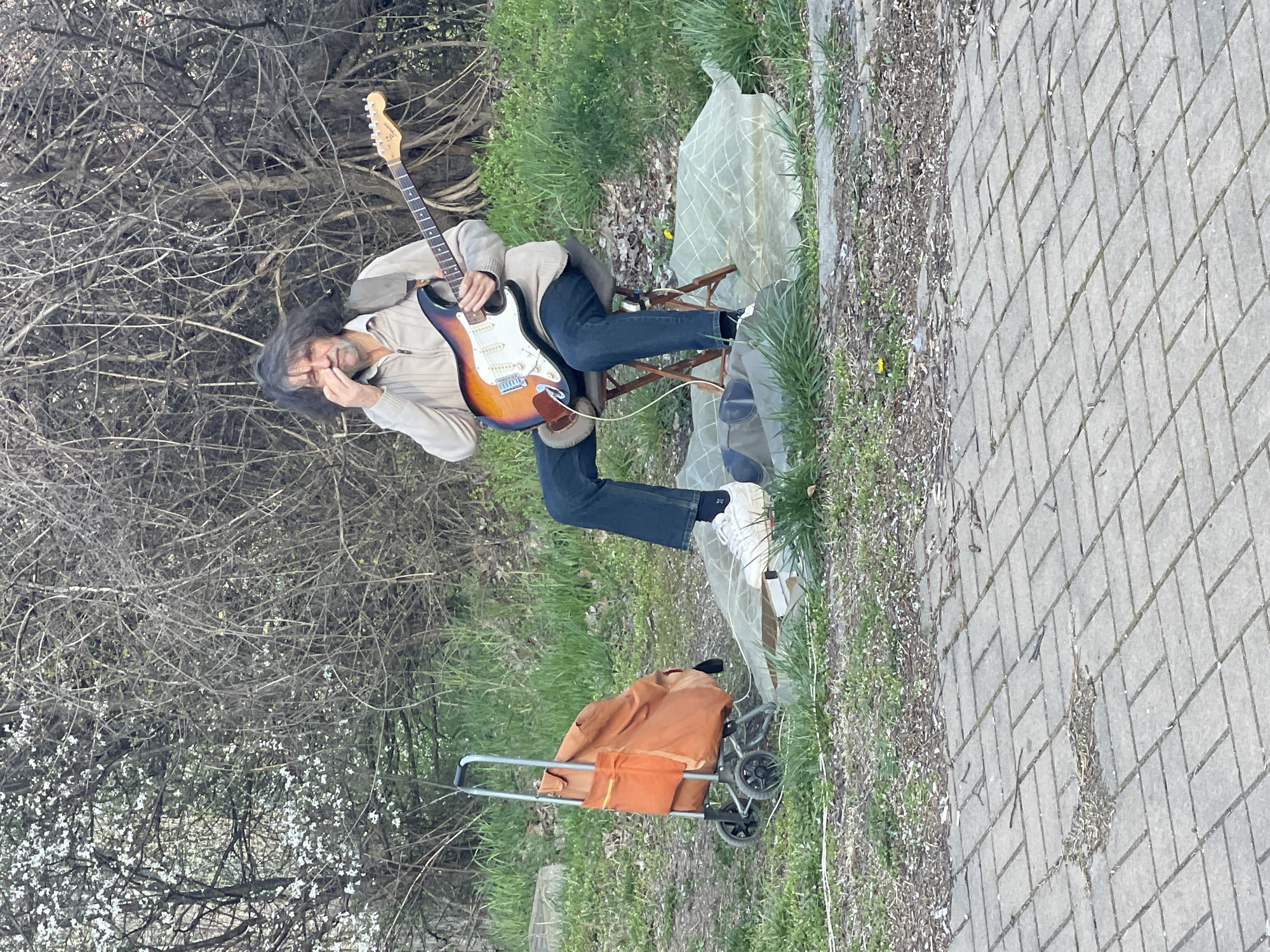A man with long dark hair and glasses sits on a folding chair in a grassy area near a sidewalk and a bare tree with some white blossoms. He wears a light brown jacket, dark blue jeans, and white sneakers. He is holding and playing an electric guitar. A shopping cart with an orange bag sits next to the chair. The location is plausibly in Sofia, Bulgaria, based on architectural styles and flora.