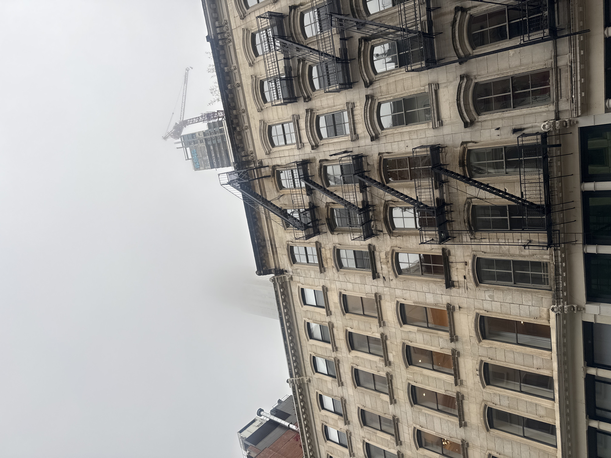 This is a low-angle, eye-level, vertical, full shot of a light-colored, multi-story building in New York City, USA, on a foggy day. The building has a classic architectural style with decorative cornices and arched windows on the upper floors, which are uniformly arranged and feature dark frames. Fire escapes, made of black metal, crisscross the facade.
In the background, a taller, modern building under construction is visible, topped with a red construction crane. The upper portions of the buildings and the sky are obscured by dense fog, creating a muted and atmospheric scene. The lower part of the building shows shops with large windows and columns. The overall impression is of an urban environment experiencing hazy weather conditions.