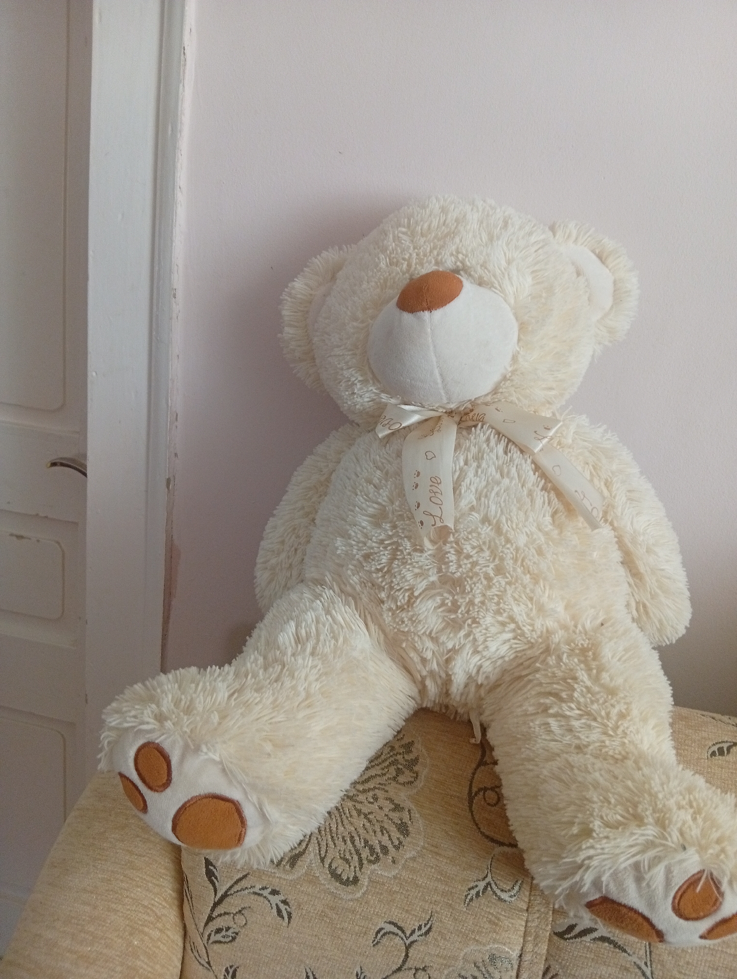 A plush, cream-colored teddy bear is positioned on a patterned sofa, leaning against a pale pink wall. The bear is facing to the left, with its legs extended forward. It wears a white ribbon with the word "Love" printed on it, tied in a bow around its neck. The bear's paws have brown paw pads. To the left of the bear is a white doorframe with a handle. The background is a plain, pale pink wall. The scene appears to be indoors, in a room within a residential building. The location is described as Veliatyno, Ukraine.
