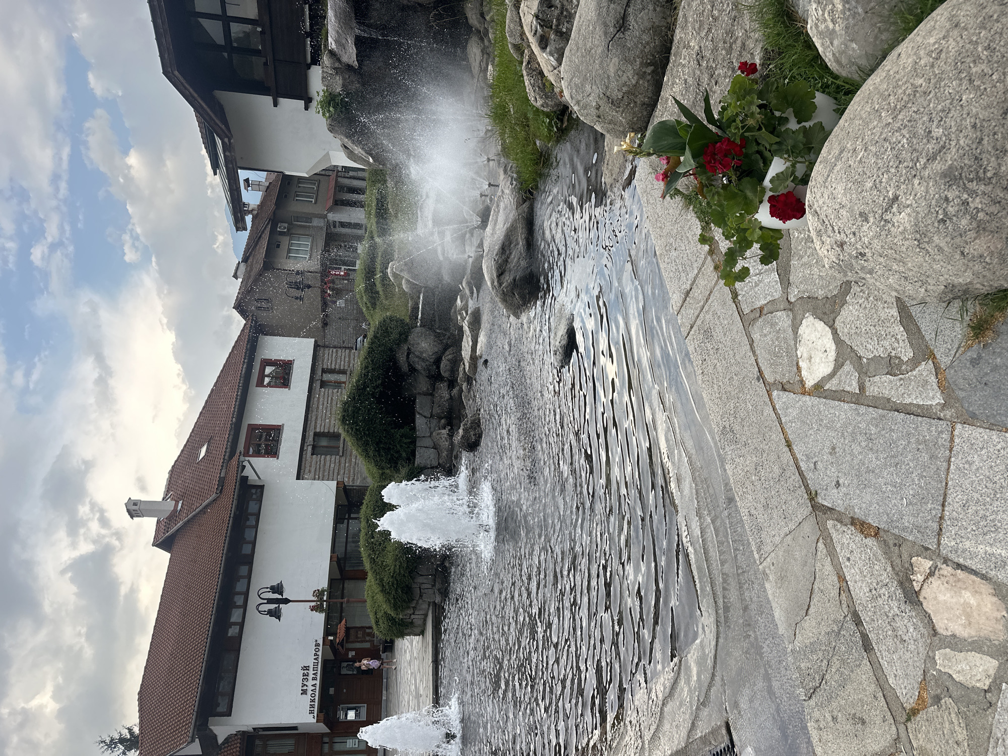 The image depicts a public space in Bansko, Bulgaria, featuring a fountain with multiple jets of water spraying upwards. The fountain is situated in a stone-lined pool, with large, rounded rocks and greenery surrounding it. In the background, traditional Bulgarian architecture is visible, including buildings with red-tiled roofs and white facades. One building has a sign that reads "МУЗЕЙ 'НИКОЛА ВАПЦАРОВ'", indicating a museum dedicated to Nikola Vaptsarov. A lamppost stands in front of this building. A person is walking in front of the museum entrance. The sky is overcast with clouds, suggesting a cloudy day. A potted plant with red flowers adds a touch of color to the foreground on the right side of the frame. The paving stones in the foreground are irregularly shaped and varied in color, indicating a rustic design.