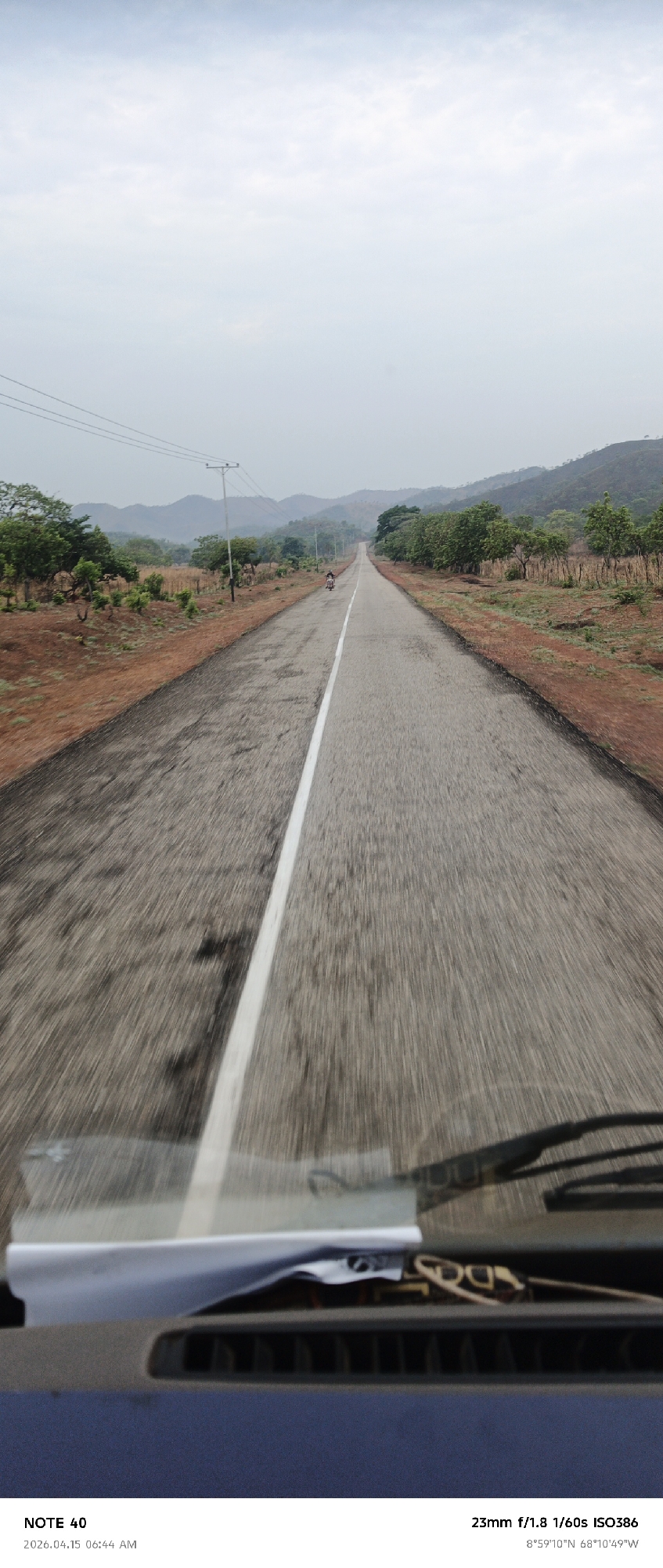 Here is a detailed description of the image:
**Scene**: The image captures a view from inside a vehicle (possibly a car or bus) driving on a straight, paved road in a rural setting. The road has a white dividing line and stretches towards the horizon.
**Environment**: The surroundings consist of scrubby vegetation and trees on both sides of the road. In the distance, there are hills or low mountains. The sky is overcast, suggesting a cloudy day.
**Objects**:
* **Vehicle Interior**: The dashboard and windshield wipers of the vehicle are visible in the foreground. There also appears to be some clutter or documents on the dashboard.
* **Road**: The asphalt road shows signs of wear, with patches and unevenness.
* **Utility Poles**: There are utility poles along the side of the road.
* **Person on a Motorcycle**: In the distance, there is a single person riding a motorcycle on the road.
**Location**: The image metadata specifies the location as approximately 8°59′10″N latitude and 68°10'49"W longitude. While that is near La Cruz, Venezuela, the exact place cannot be pinpointed just from this GPS coordinate alone. Additional details in the image, such as landmarks or road signs, would be needed to confirm.