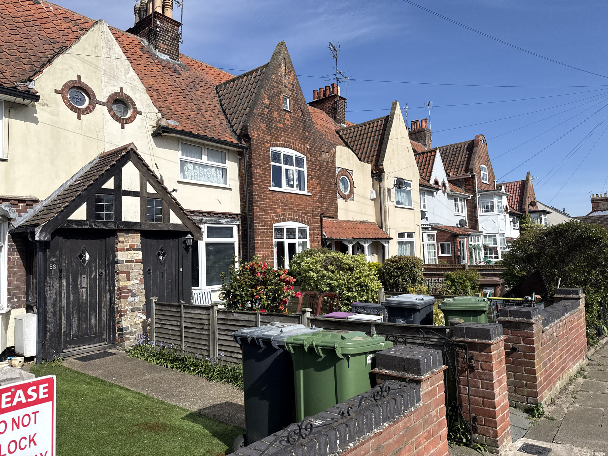 The image shows a row of terraced houses on a street in Gorleston-on-Sea, United Kingdom. The houses are primarily constructed of brick and feature a variety of architectural styles, including gabled roofs, round windows, and half-timbered detailing. In front of the houses are waste bins and low brick walls with ironwork. A patch of artificial turf is visible near a red sign that reads "EASE DO NOT LOCK". The sky is blue with no clouds.