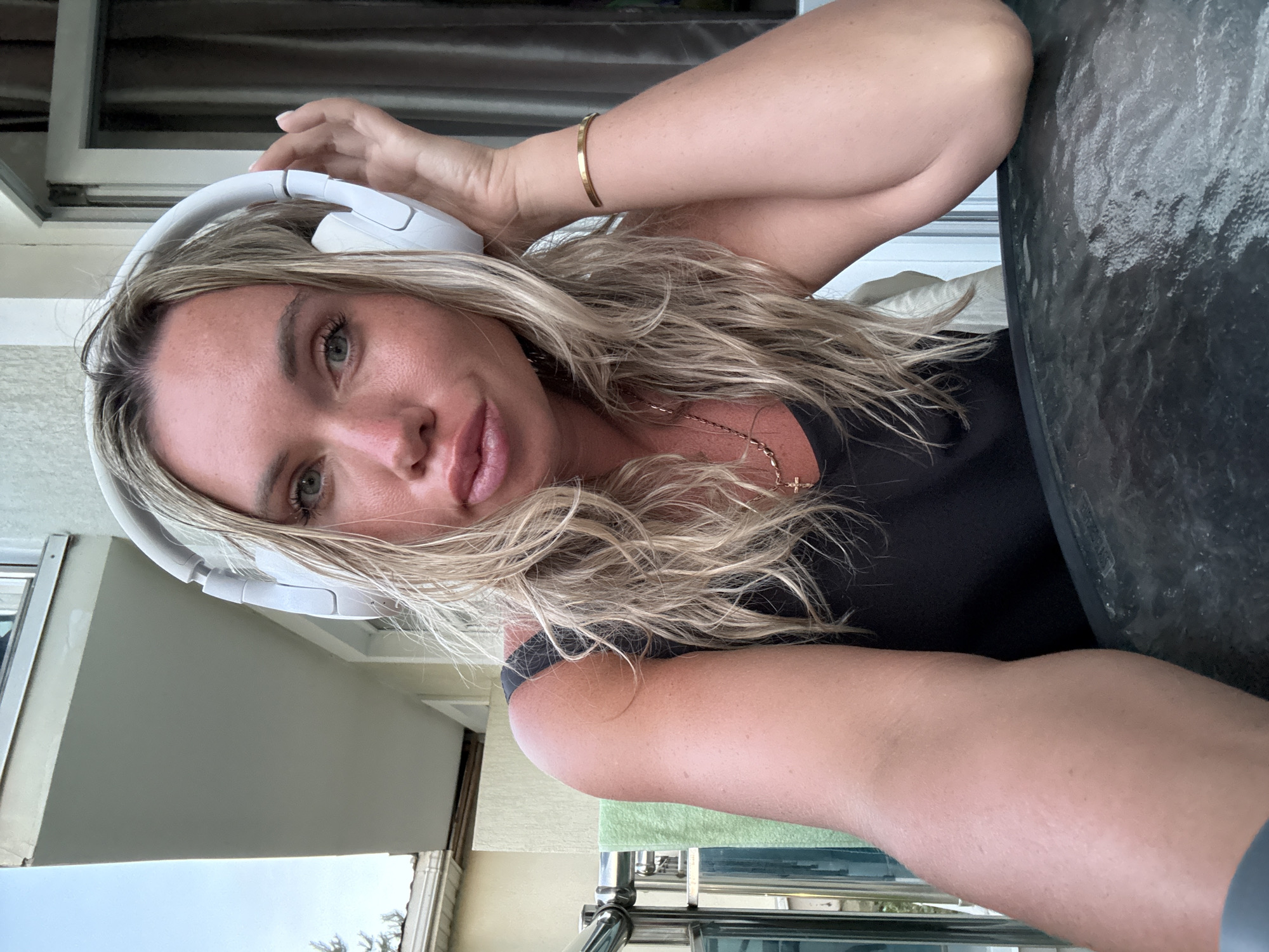 In Anapa, Russia, a blonde woman wearing white headphones and a black tank top sits at a glass-topped table outdoors, with a balcony railing and a building in the background. She is looking at the camera with her hand resting on her headphones. Her blonde hair is styled loosely, and she is wearing a gold necklace with a cross pendant and a gold bracelet on her right wrist. The lighting suggests it is daytime.