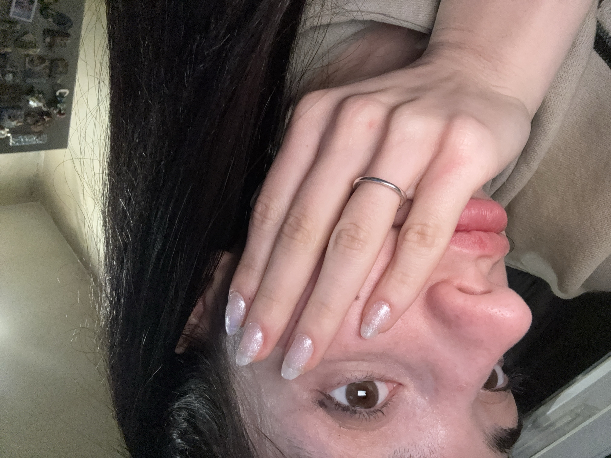 The image shows a person with long, dark hair partially covering their face. The person's right hand, adorned with a silver ring on the ring finger and sparkly, light-colored nail polish on the elongated nails, is positioned over their face. The person has light skin, brown eyes, and a pink nose. The background is blurry but appears to include a light-colored surface and a beige fabric. There is no information to determine the location of the image in Sofia, Bulgaria.