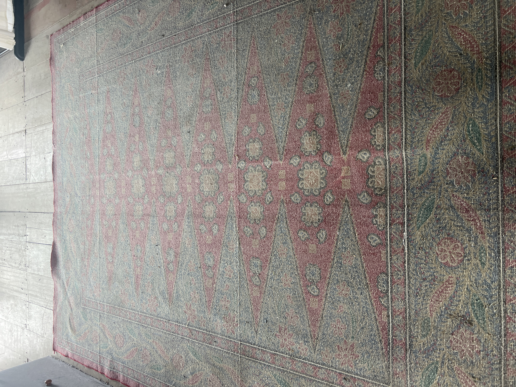 An overhead shot shows a worn, oriental rug with a red, blue, and green pattern spread across a light-colored wooden floor. The rug's central design features repeating diamond shapes, outlined in blue and filled with red and floral motifs. The rug appears to be in a room in Kuta Selatan, Indonesia, likely a residential or commercial space, given the presence of wooden floorboards and what appears to be a portion of a wall or skirting board on the left edge of the image. There are no people or animals visible in the scene.