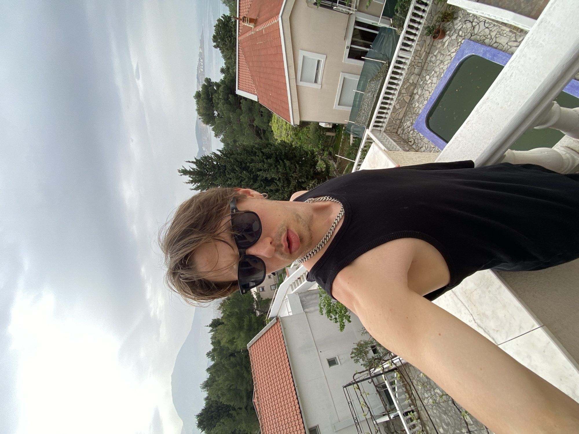 A selfie photograph shows a person with sunglasses and a black tank top, wearing a silver chain, on a balcony in Šušanj, Montenegro. The image includes a partial view of the person's arm and upper torso. The background reveals a cloudy sky, green trees, a house with a red tile roof, and a swimming pool with discolored water. The balcony has a white railing. The photo appears to be taken from an elevated position, looking downward.