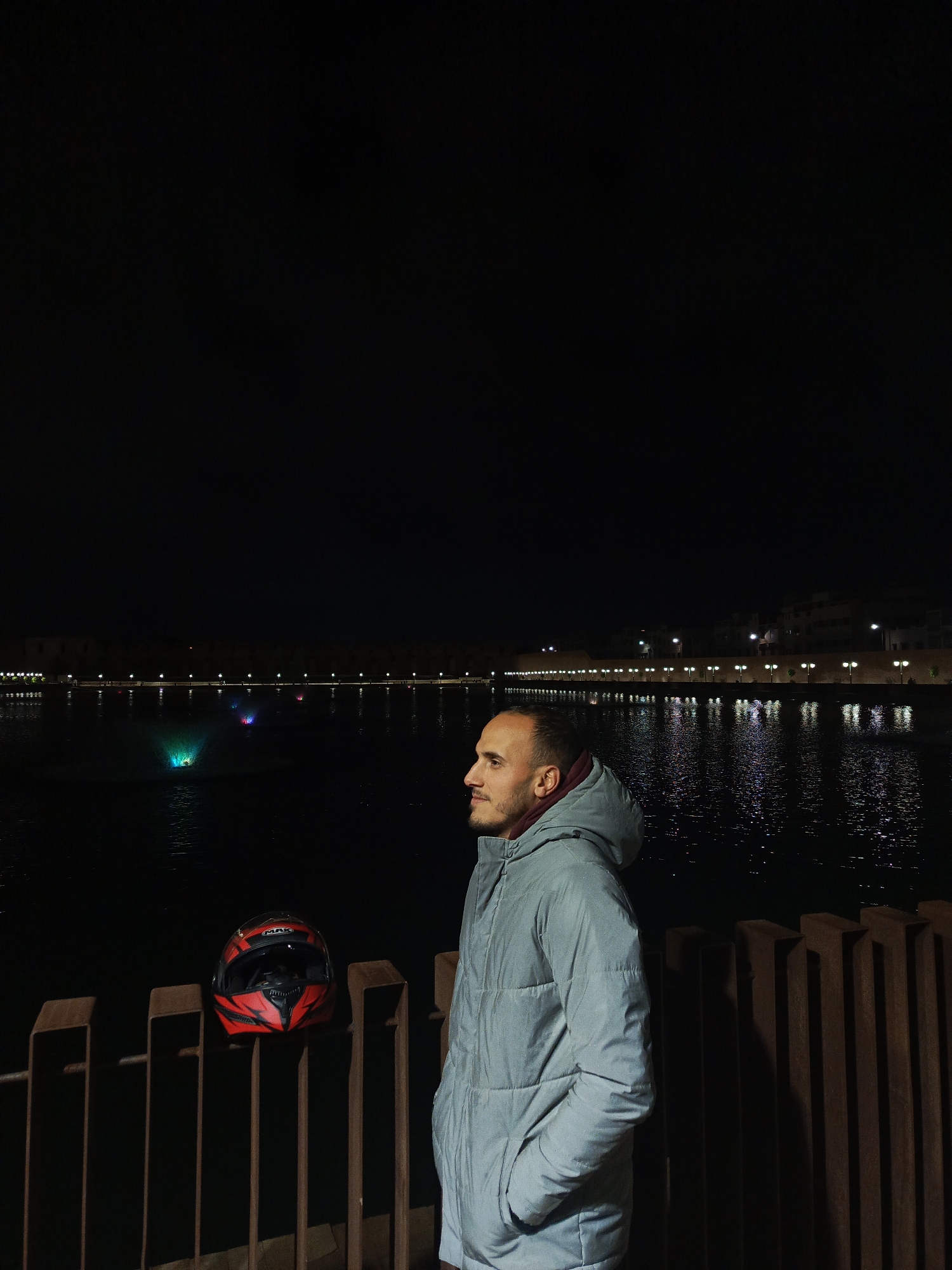 A man stands at night in Al Machouar – Stinia, Morocco, near a body of water with illuminated fountains and building lights reflecting on the water's surface. He wears a light-colored hooded jacket and is positioned near a fence with a red and black motorcycle helmet resting on it. The background is dark, with the lights from the buildings and fountains providing illumination.