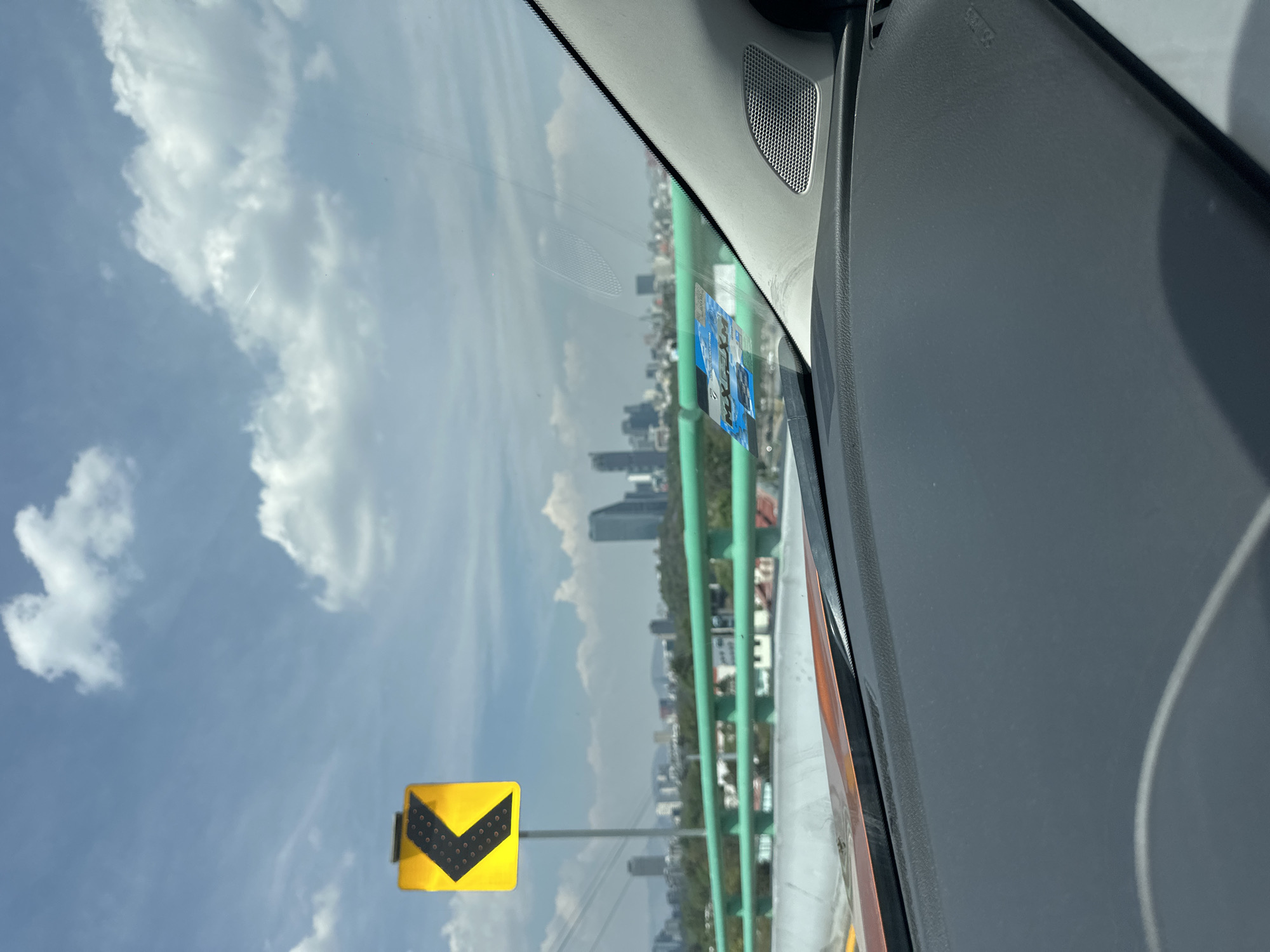 The photograph, taken from inside a vehicle, captures a view through the windshield. The exterior scene features a bright blue sky with scattered cumulus clouds. A yellow directional sign with a black chevron pointing to the left is prominently visible, suggesting an upcoming curve or turn in the road. Beyond the sign and the road, a cityscape is visible, with tall buildings and green foliage. The green structure appears to be an elevated freeway. The registration stickers are visible on the windshield. Given the presence of an elevated freeway and the style of buildings, this photograph was likely taken in Mexico City, Mexico. The interior of the car includes a gray dashboard and speaker grill.