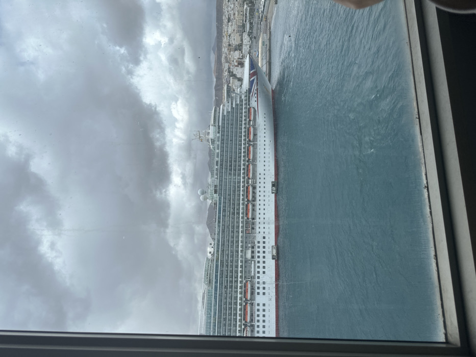 The image presents an exterior view through a window, showcasing the harbor of Puerto del Rosario, Spain, under an overcast sky. A large cruise ship, identified as "AZURA" by text on its bow, dominates the midground. The ship features a white hull with a red stripe along the waterline, blue-green balcony decks, and numerous orange lifeboats secured along its side. Its bow is oriented towards the right, displaying a Union Jack flag design. The harbor water is dark blue-green with visible surface ripples. To the right of the ship, a segment of the city of Puerto del Rosario is visible, comprising tightly packed light-colored buildings. Low, arid, reddish-brown hills rise in the distant background behind the cityscape. The sky is entirely covered by dense, grey clouds. The foreground includes a grey window frame, with minor smudges or water marks visible on the glass surface. No individuals are clearly discernible within the scene.