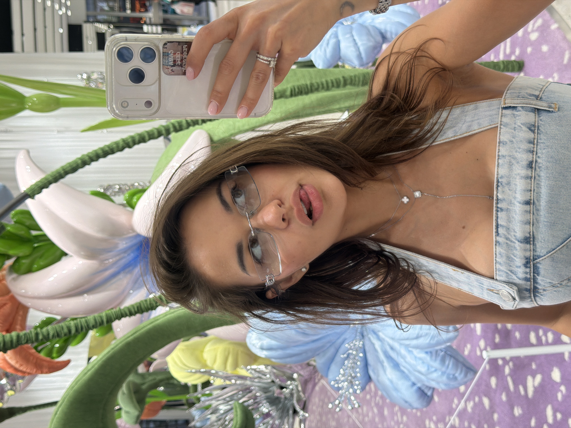 A woman is taking a selfie in what appears to be a brightly decorated indoor space. She is wearing a light blue denim overall top and glasses, and has a silver necklace with flower-like pendants. She holds an iPhone in her hand, showing a partial view of the phone's back and camera lenses. She has brown hair.
Behind her, there are large artificial flowers in colors like white, blue, yellow, orange, and green. The floor has a purple and white spotted pattern.
The city of Dongcheng District, China is not explicitly shown.