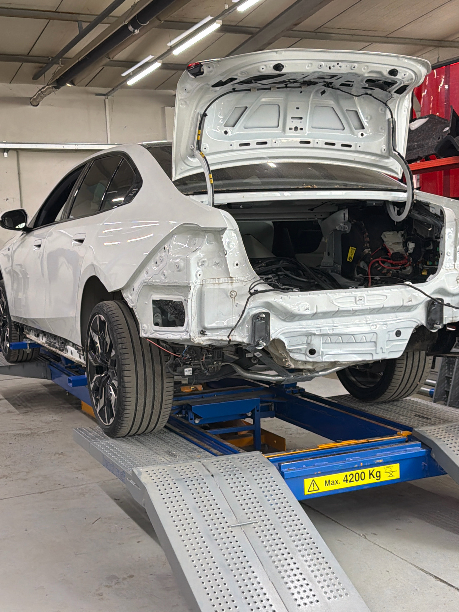 An image shows a white sedan being worked on in a garage. The car is on a hydraulic lift, with its trunk open, revealing exposed internal components. The rear bumper and surrounding bodywork are removed, exposing the car's chassis and wiring. The garage appears to be a professional auto repair shop, with tools and equipment visible in the background. There is a yellow warning sign on the lift that reads "Max. 4200 Kg". The overall scene suggests that the car is undergoing significant repairs or modifications, possibly related to collision damage or structural work. The image was taken in the city of Burgas, Bulgaria.