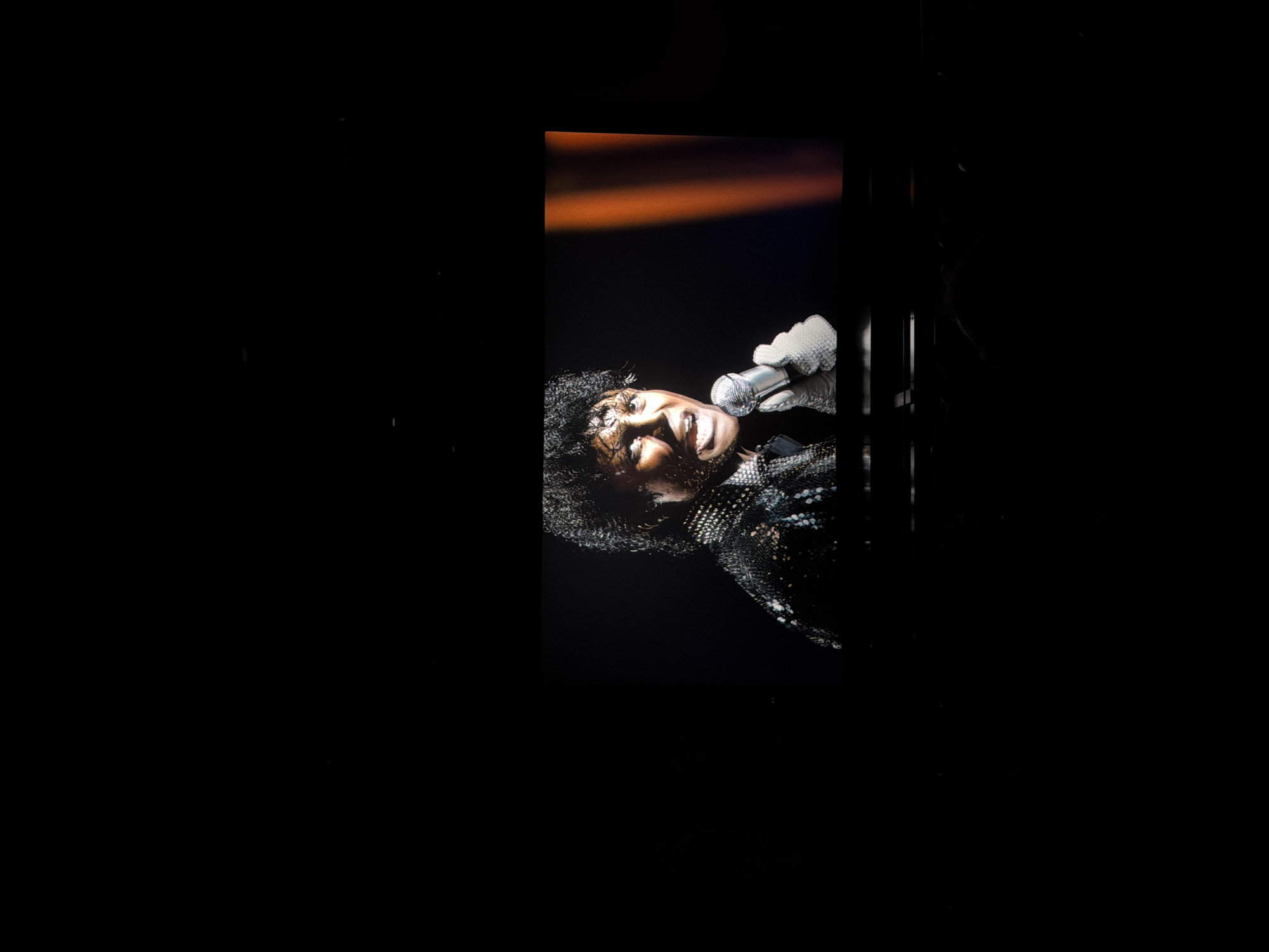 The image depicts a performer, strongly resembling Michael Jackson, captured mid-performance. The subject is seen from the chest up, positioned on the right side of the frame, within a brightly lit, vertical rectangular area against an otherwise dark background. Their head is tilted upwards and to the left, with an intense expression, mouth open as if singing. They hold a metallic microphone in their right hand, which is adorned with a distinctive white, textured glove.
The performer is wearing a dark, sequined or sparkly jacket/top, catching the light. Their hair is dark and curly. Above and behind the performer's head, an orange spotlight or stage light casts a warm glow across the dark background. The overall scene suggests a live concert or stage performance, likely indoors or at night, given the stark contrast between the illuminated subject and the surrounding darkness. No text is visible in the image.