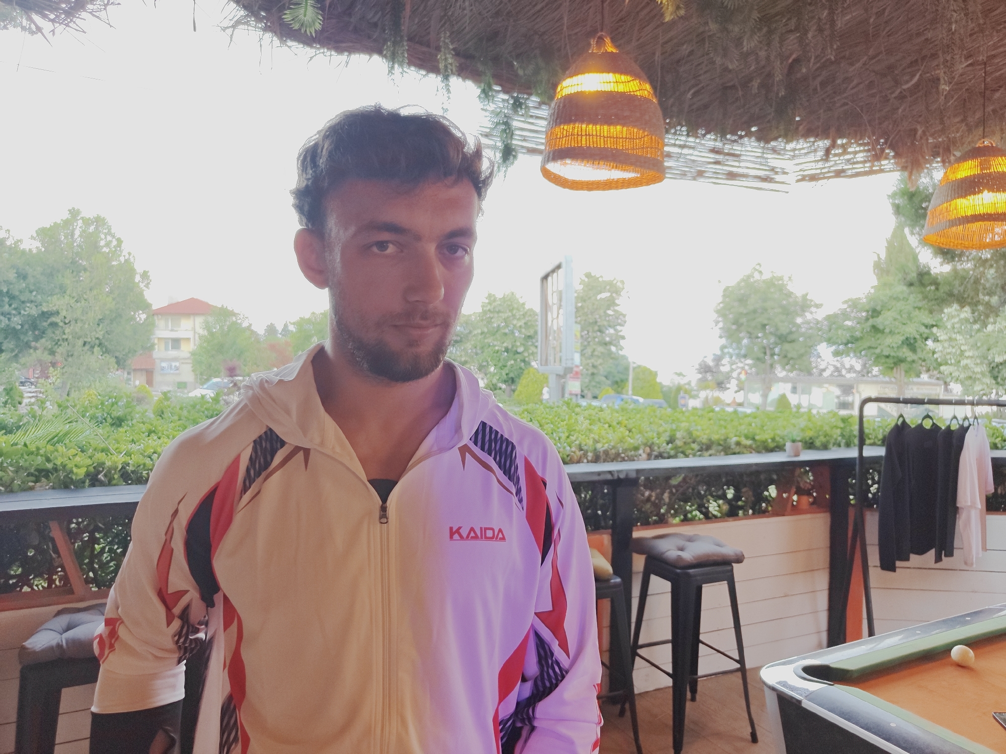 A man with dark hair and a beard is standing outdoors, wearing a white and pink hooded jacket with red and black accents. He is looking at the camera. In the background, there are trees, buildings, and hanging lamps. There is also a bar, stools, a clothing rack with shirts, and a pool table with a ball. The scene appears to be an outdoor seating area of a establishment in Tsarevo, Bulgaria.