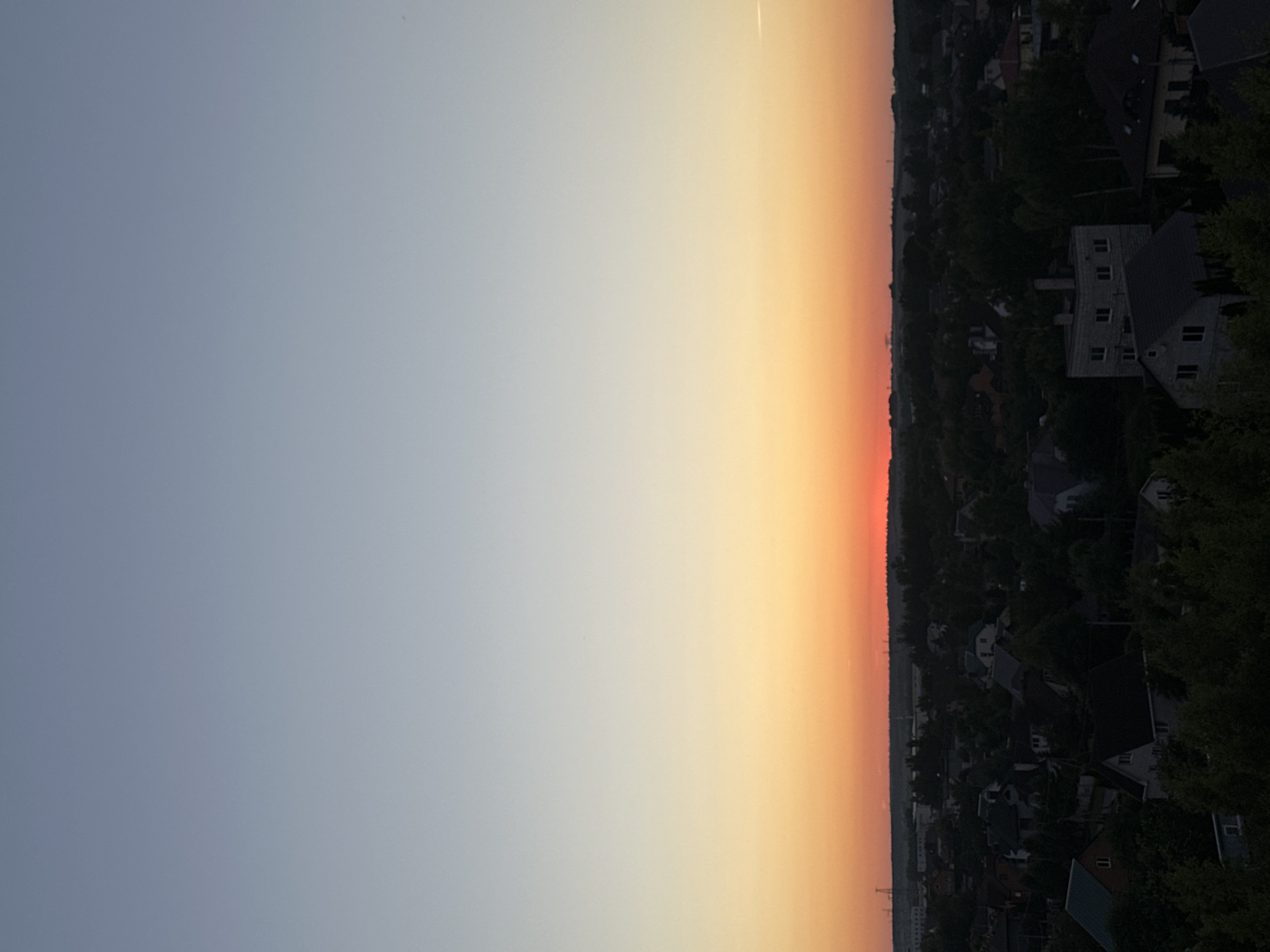 The image depicts a panoramic view of a residential area at dusk, likely within the Шчомысліцкі сельскі Савет in Belarus. The sky transitions from a pale blue at the top to a vibrant orange hue near the horizon, indicating the setting sun. Below the sky, a dense canopy of trees and rooftops of houses cover the landscape. The houses are varied in design and color, with some featuring dark roofs and others lighter facades. The overall impression is one of a quiet, suburban or semi-rural environment bathed in the soft light of twilight. There are no people visible in the frame.