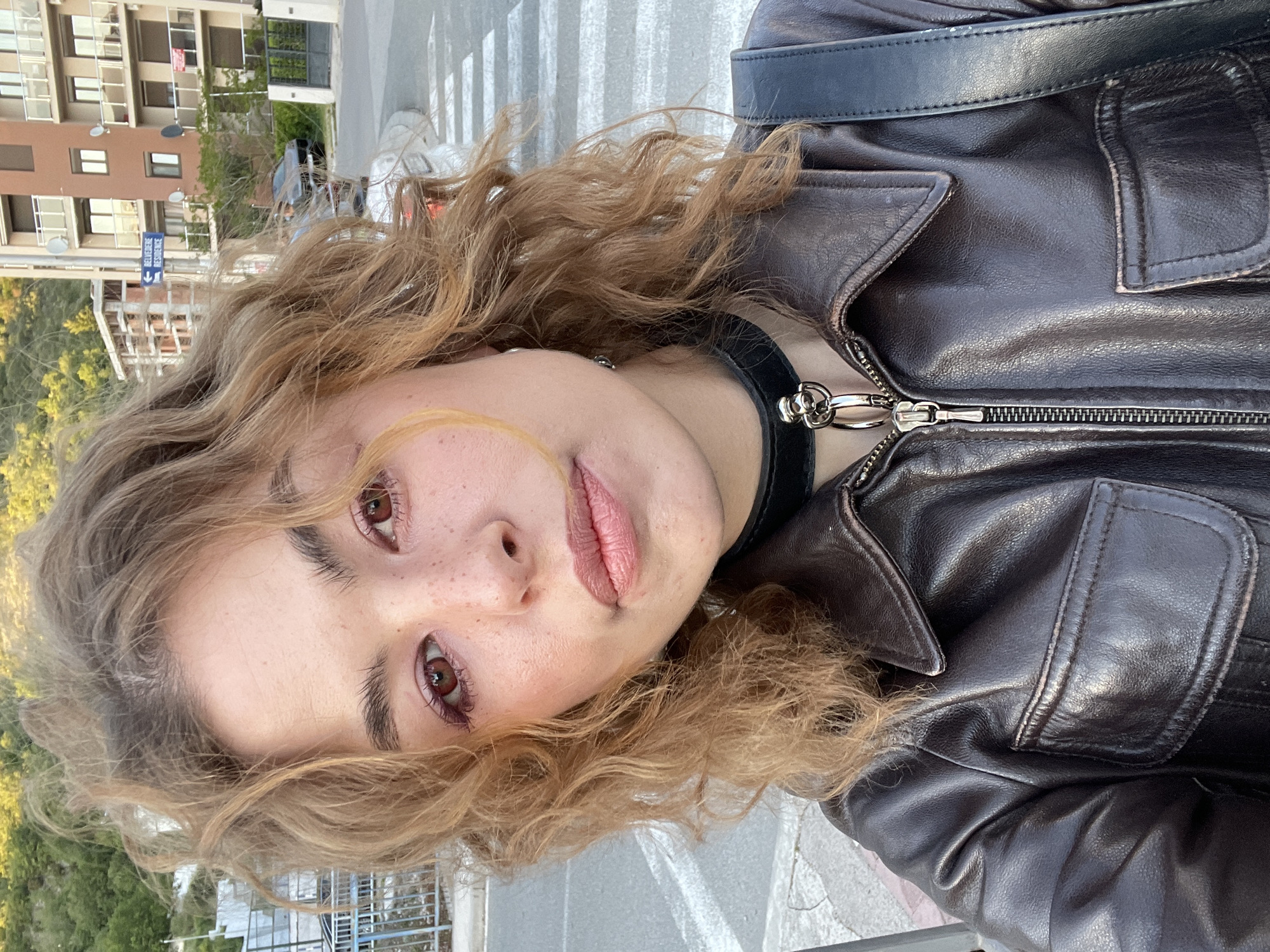 The image is a close-up, selfie-style portrait of a young person, rotated ninety degrees to the left. The subject has light brown, wavy hair that frames their face. They are looking directly at the camera with a neutral to slightly contemplative expression, showcasing one dark brown eye, part of their nose, and soft pink lips. Freckles are visible across their nose and cheeks. The person is wearing a dark brown or black leather jacket, which features a prominent silver zipper running down the front. A black choker with a metallic silver clasp is visible around their neck.
The background depicts an outdoor urban or suburban setting. Several multi-story residential buildings with balconies and varied facades are visible behind the subject. A street with a white crosswalk is discernible in the mid-ground, along with glimpses of vehicles. Green trees and foliage are present on the left side of the frame, providing a natural element. A blue directional sign with white text is clearly visible on a pole in the background. The sign reads "BELVEDERE RESIDENCE" with an arrow pointing left, indicating the direction. There is additional, less legible text below "RESIDENCE." The scene is illuminated by natural daylight, suggesting it is daytime. The location context provided indicates this scene takes place in Boreti, Montenegro.