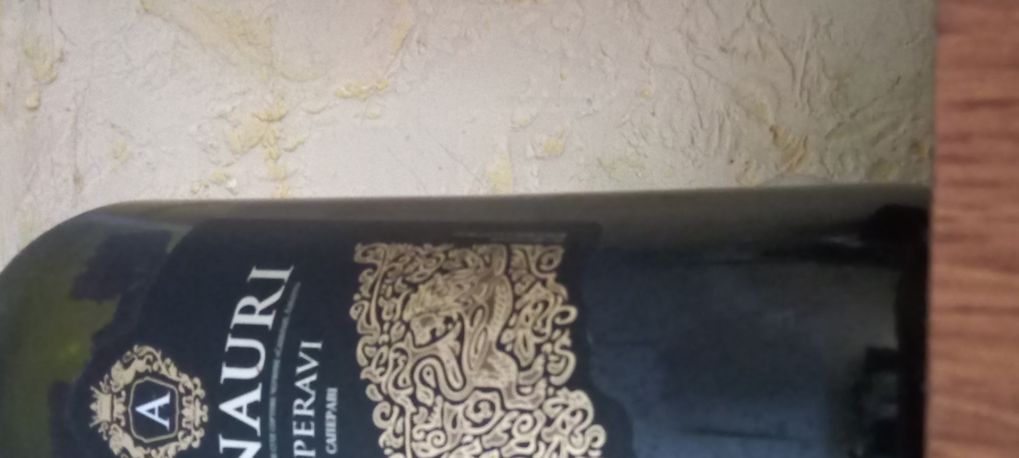 A dark-colored wine bottle, possibly red wine, stands on a wooden surface against a textured, off-white wall. The bottle has a black label with gold lettering and a gold decorative emblem featuring a crown and the letter "A". The text on the label is partially visible, reading "NAURI" and "PERAVI". Below this, in smaller font, it says "CABERNET". A gold ornamental design, possibly a stylized lion or griffin, is printed on the lower part of the label. The background wall appears to be plaster with an uneven, textured finish, possibly wallpaper. The lighting suggests an indoor setting. The image is cropped to focus on the bottle and its label. There are no people or discernible activity in the scene. There is no information present in the image to indicate its location in Kryvyi Rih, Ukraine.
