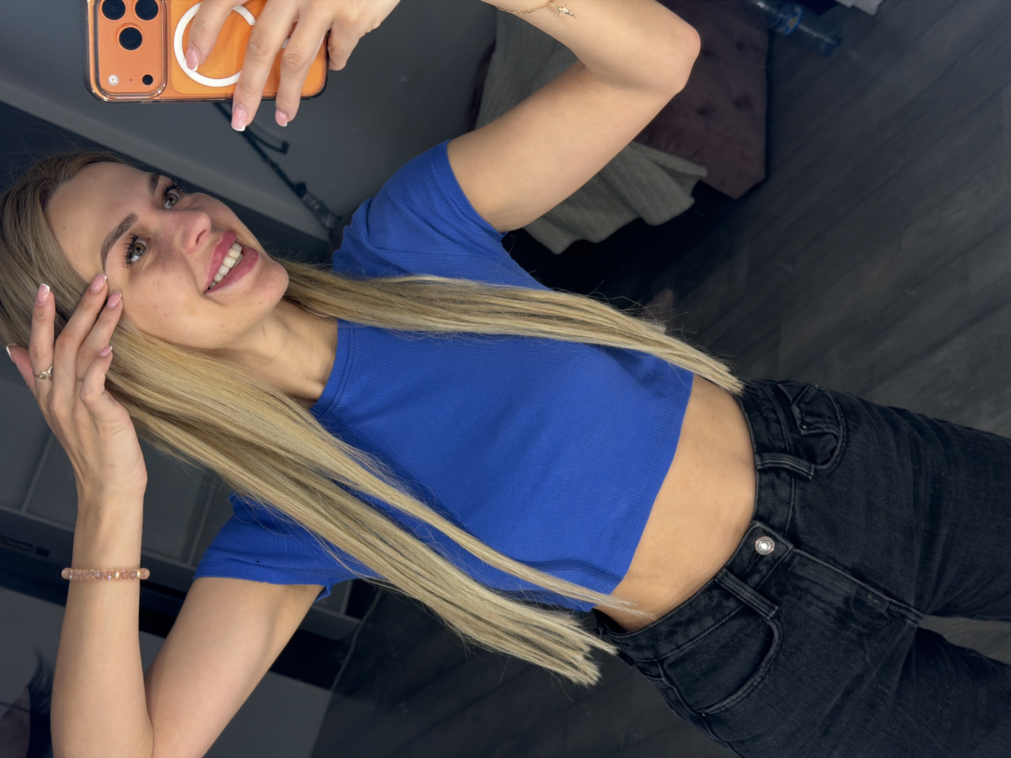 A young woman with blonde hair is taking a selfie in a room. She is wearing a blue crop top and black jeans. She has a bracelet on her left wrist and a ring on her right index finger. She is smiling and holding her phone up to take the picture. The room appears to be a bedroom or dressing area, with a bed and a closet visible in the background. The flooring is a dark wood. There is no indication of the specific location within Kyiv, Ukraine.