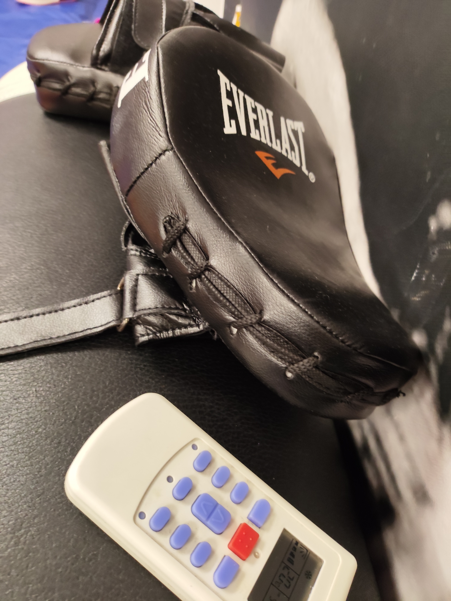 The image shows a pair of black Everlast boxing mitts resting on a black textured surface. In the foreground, a white remote control with blue and red buttons is partially visible. The remote has a small digital display showing some symbols and numbers, including "-02". The background shows a blurry image of a white abstract shape or pattern on a dark surface. There are no people visible in the image. The scene appears to be indoors, possibly in a gym or a home setting, but there are no specific visual cues that would allow for determination of its location within the city of Krasnodar, Russia.