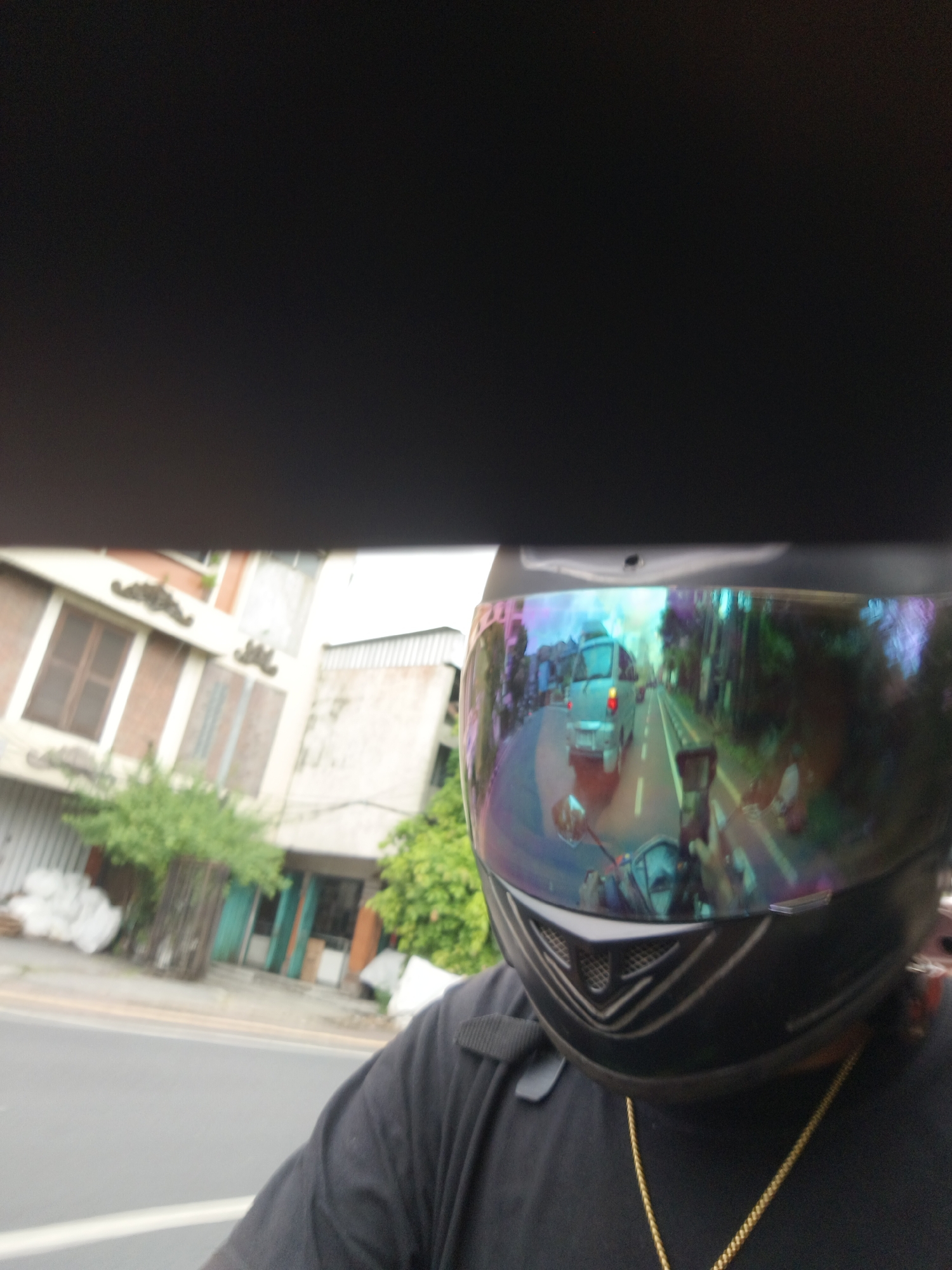 The image shows a first-person perspective of someone riding a motorcycle in Denpasar, Indonesia. The rider is wearing a black helmet with a mirrored visor, which reflects a street scene with a silver van driving ahead. Buildings line the street, with one on the left appearing to be a two-story structure with brick accents and brown-framed windows. Trees and foliage are visible on the right side of the road. The rider is wearing a black t-shirt and a gold chain necklace. The overall impression is a snapshot of daily life and transportation in a Southeast Asian city.