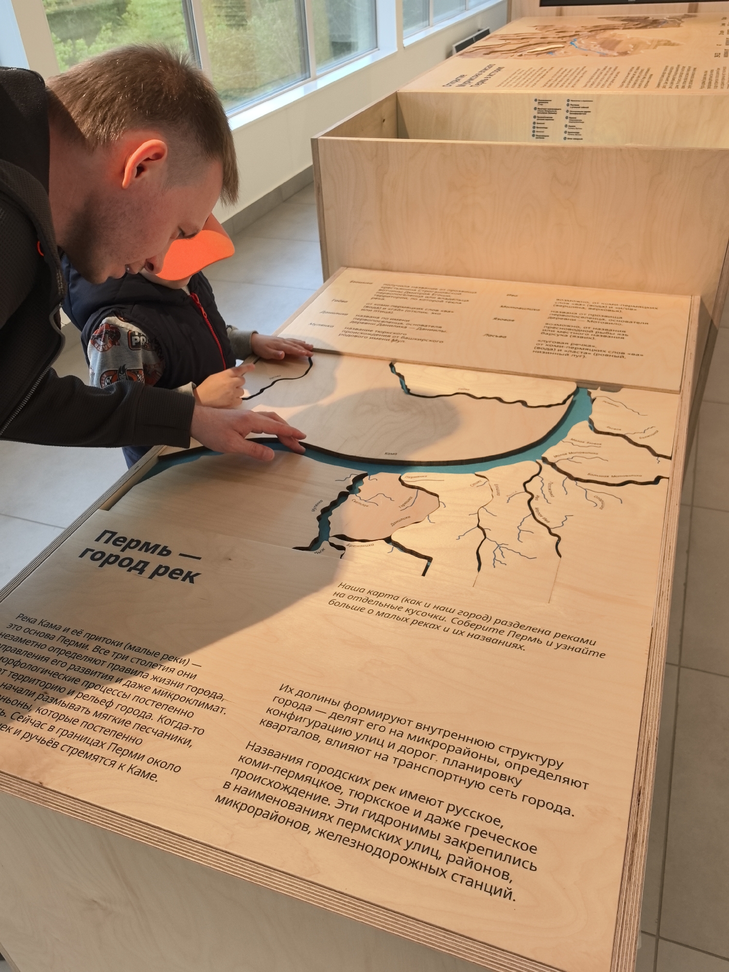 An adult male and a young child are interacting with an educational exhibit in an indoor setting, likely a museum or science center, in Perm, Russia.
The adult male, appearing to be in his 30s or 40s with short, light brown hair and wearing a dark, zippered jacket, leans over the exhibit table. His right hand rests on a wooden piece of the interactive map, while he observes the child. The child, approximately 2-5 years old, wears an orange cap, a dark blue vest, and a light-colored long-sleeve shirt with a print. Both of the child's hands are placed on separate wooden map pieces, actively engaging with the exhibit.
The central subject is a large, light-colored wooden interactive map display. It depicts a river system, specifically the Kama River and its tributaries, which form the geography of Perm. The map is composed of numerous individual, removable wooden landmass pieces, separated by carved channels filled with a blue material representing the rivers. Visible labels on the wooden pieces include "КАМА," "Данилиха," "Мотовилиха," "Стикс," and "Мулянка," among others, all written in Russian.
Integrated into the exhibit are several Russian text panels. A prominent heading to the left reads "Пермь — город рек" (Perm — city of rivers). This panel describes how the Kama River and its tributaries form the foundation of Perm, influencing its life, development, microclimate, and terrain. Another panel to the right explains how the river valleys shape the city's internal structure, defining micro-districts, street layouts, and transportation networks. It also details the diverse origins of city river names—Russian, Komi-Permyak, Turkic, and Greek—and how these hydronyms are preserved in the names of streets, districts, and railway stations. Further text panels are visible on the upper part of the exhibit, including a partially visible title "Открытие территории: жизнь и история" (Discovery of the territory: life and history).
The exhibit is constructed within a system of light wooden frames. In the background, a large window provides natural light, revealing green foliage and trees outside. The room is brightly illuminated.