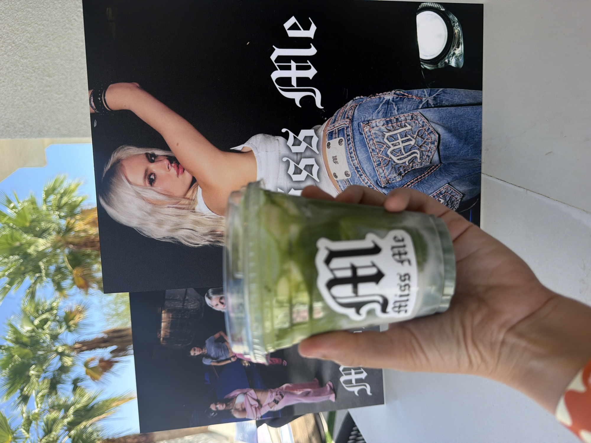 The photograph depicts a hand holding a cup with the words "Miss Me" displayed on the label, partially obscuring a promotional poster of a woman. The backdrop includes a sunny outdoor scene with palm trees, suggestive of a location with a warm climate. Based on the environment and the context of "Miss Me" likely relating to the Coachella music festival, it's reasonable to assume the photo was taken in Indio, United States of America.