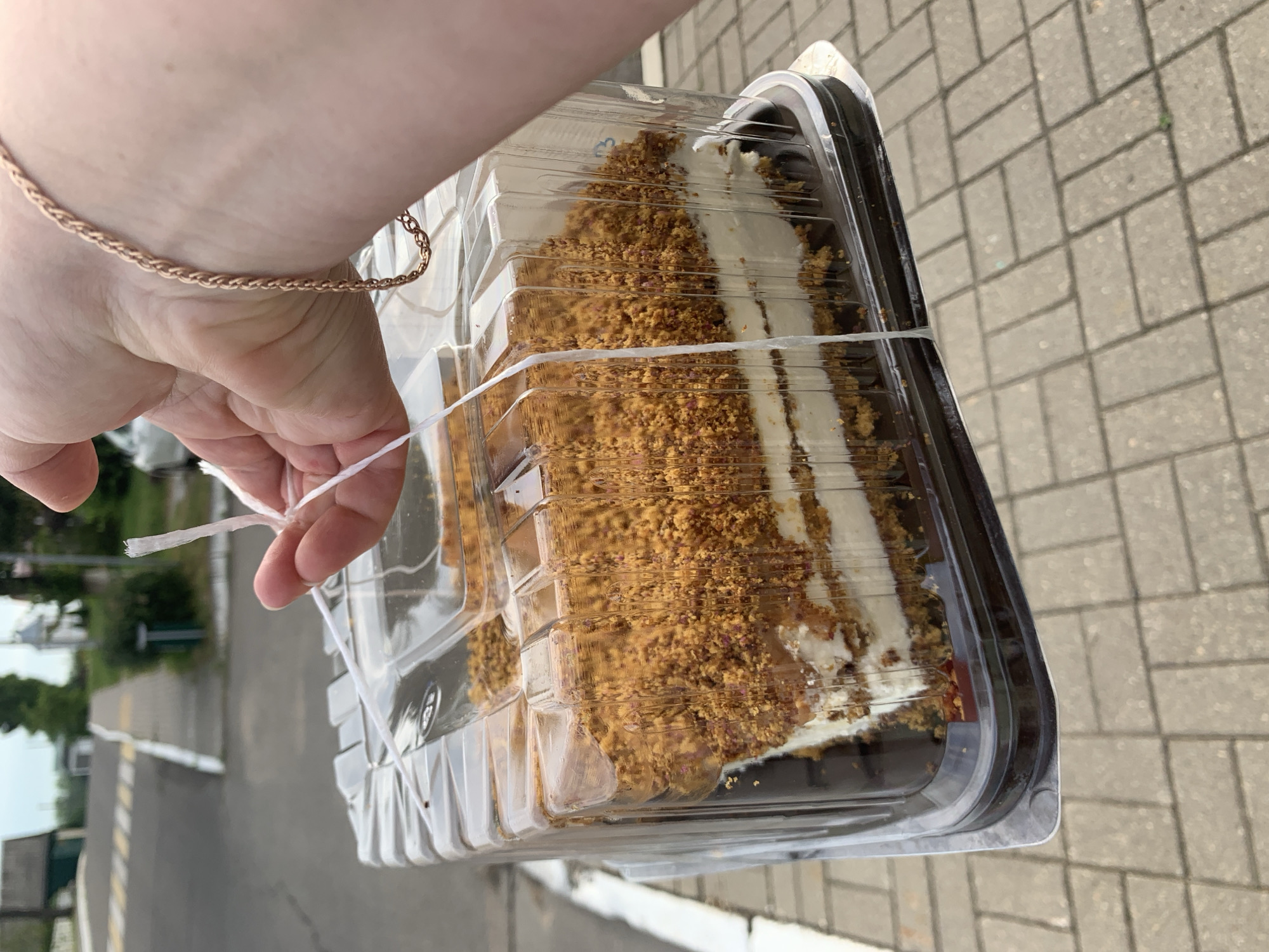 A person's hand, adorned with a braided rose gold bracelet, is holding a clear plastic container with a slice of cake inside. The cake appears to be a layered dessert with white cream filling between layers of cake. The exterior of the cake is covered in crumbled brown material, possibly nuts or cake crumbs. The container is secured with white string, which the person is holding. The background suggests an outdoor urban environment. The scene takes place on a paved sidewalk with brick-like patterns, and a road with painted lines and a pedestrian crossing is visible in the distance. Buildings and greenery can be seen further back. The weather appears to be overcast. The location is identified as the city of Kapyĺ, Belarus.