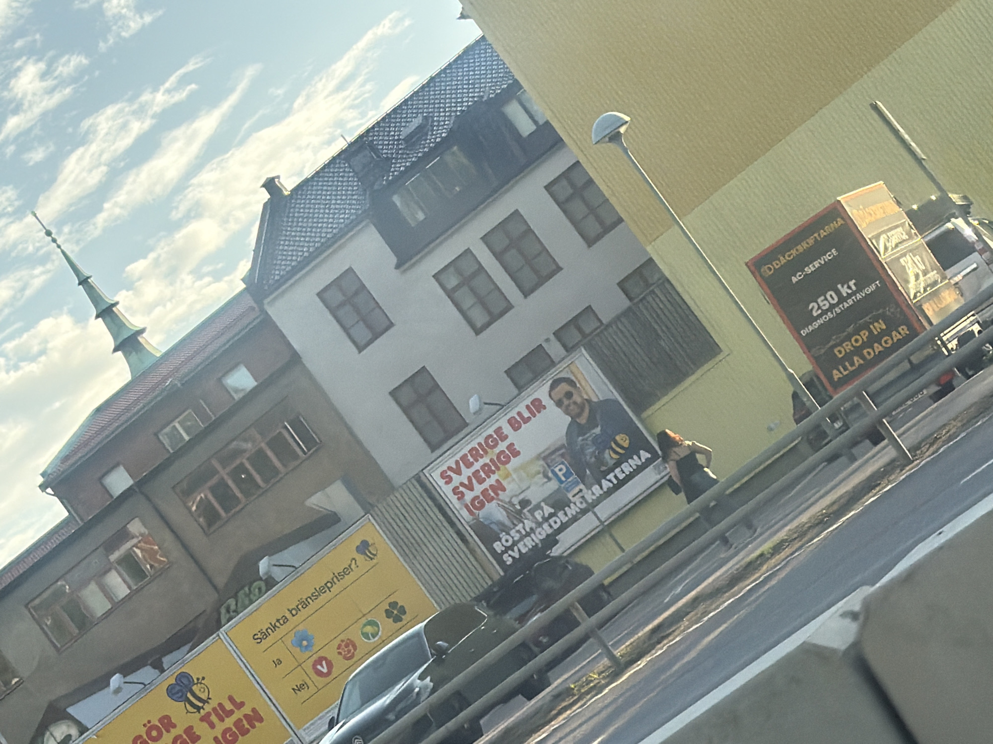 The image depicts an outdoor scene with a slightly tilted perspective, capturing a street view in Gothenburg, Sweden. In the foreground, a blurred highway or road is visible with a concrete barrier. Beyond the barrier, a woman with long, dark hair is standing near a metal railing, appearing to be looking away from the camera. To the left, a billboard displays "SVERIGE BLIR SVERIGE IGEN" with a picture of a man and a bee logo. To the right, another billboard advertises "DÄCKSKIFTARMA AC-SERVICE" and "250 kr DROP IN ALLA DAGAR". In the background, several buildings are visible, including one with a prominent spire. The sky is partly cloudy.