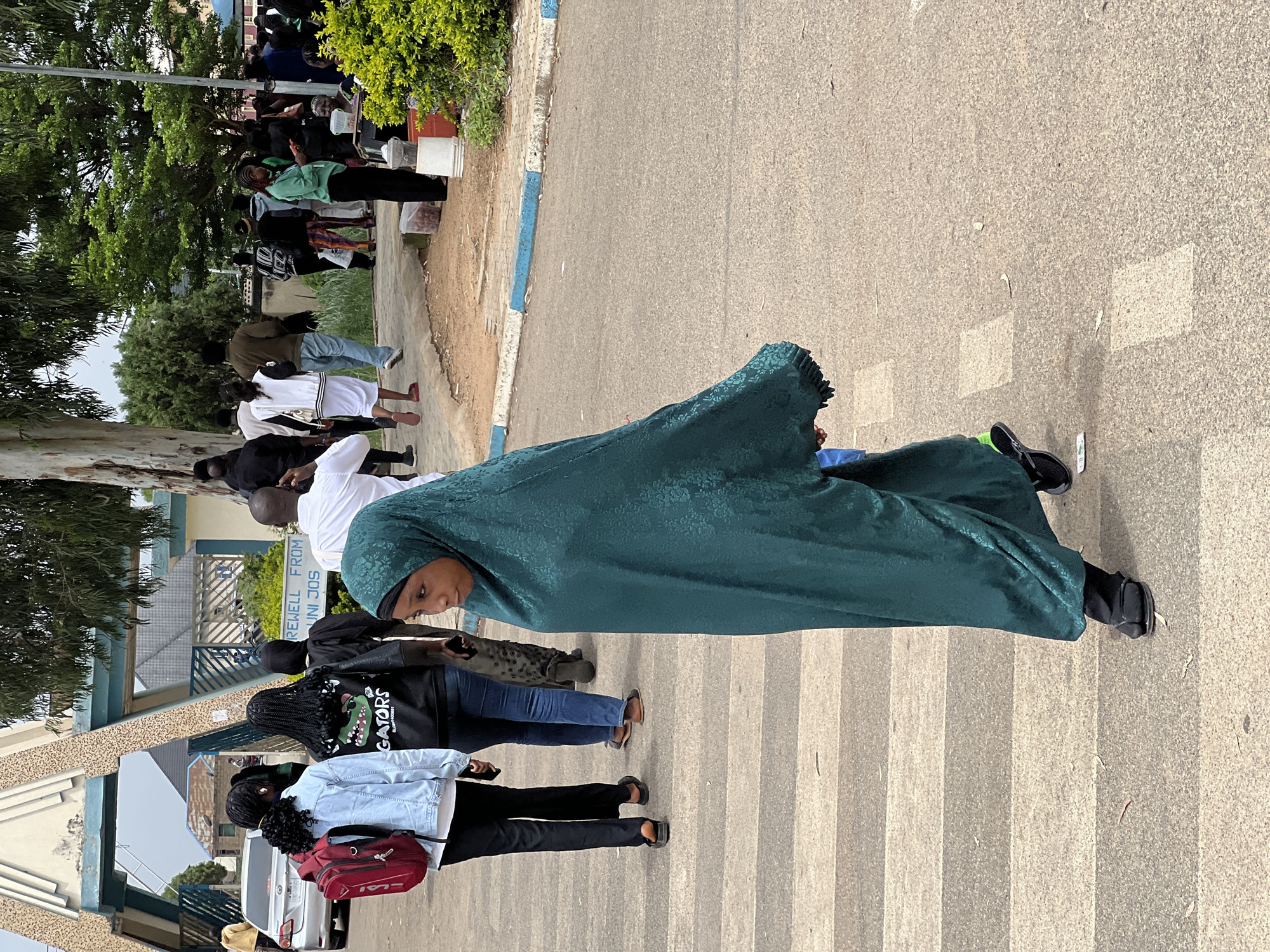 The image shows a group of people, predominantly young adults, at a pedestrian crossing. A young girl wearing a green hijab and matching dress is walking across the zebra crossing in the foreground, looking to the right. She is wearing black sandals. Behind her, several other individuals are also crossing or walking on the pavement. To the left, two young women are walking away from the camera, one in a light blue denim jacket and a red backpack, and the other in a black jacket with a graphic print. Further in the background, more people are visible, some walking towards an archway entrance that appears to be an official gateway, possibly to an institution. A silver car is parked to the left of the entrance. Signs are visible above the archway, with text that includes "FAREWELL FROM" and "UNI JOS", indicating the location is the University of Jos, Nigeria. The environment appears to be an outdoor, paved area with some trees and greenery. The overall scene suggests a departure or farewell event.