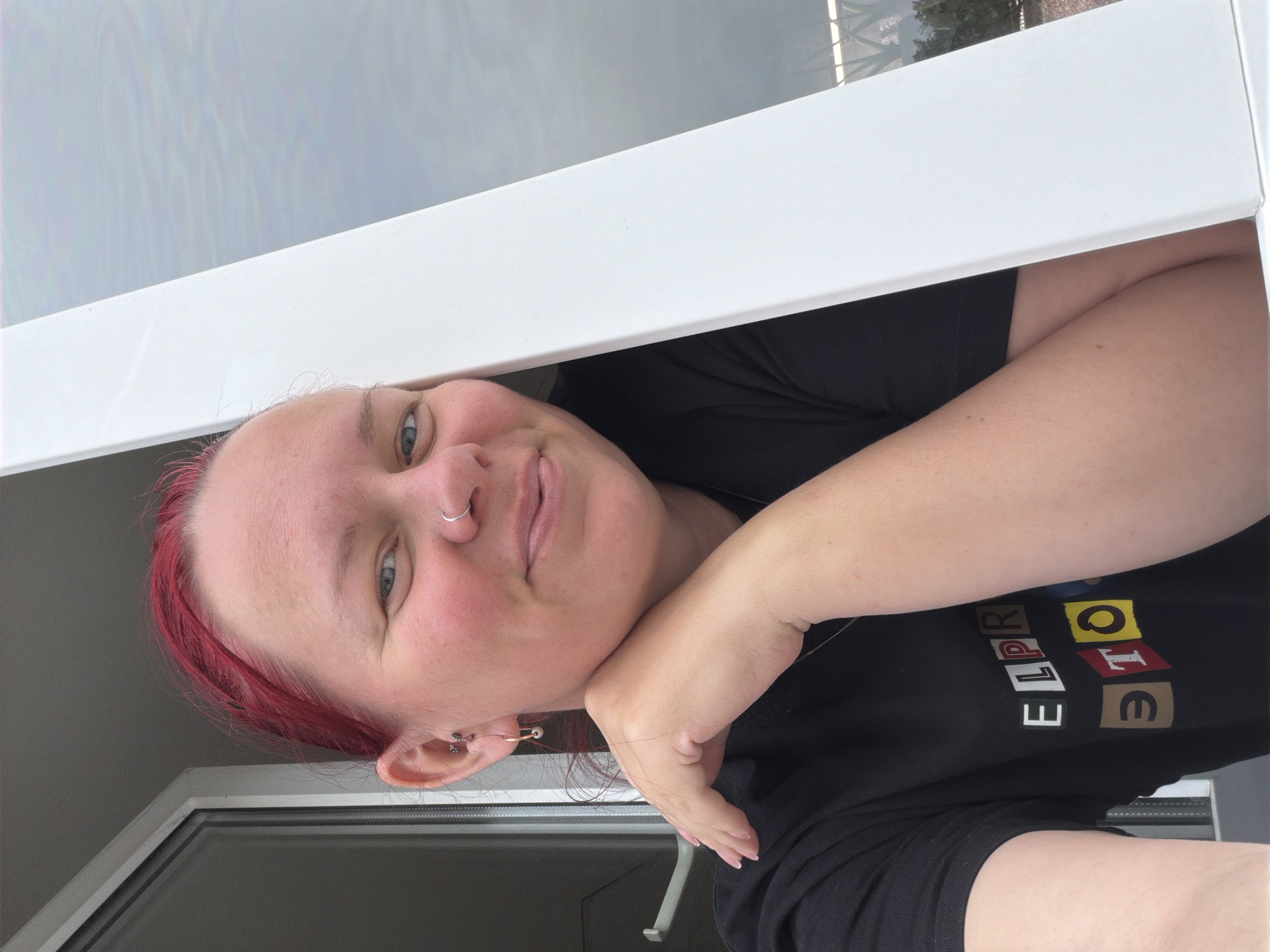 A person with reddish-purple hair and blue eyes looks into the camera. The person is wearing a black t-shirt with multicolored letters on blocks. The person has multiple piercings in their left ear, and a nose ring. They are resting their head on their hand, and looking out of a white window. The location is Chaikovsky, Russia.