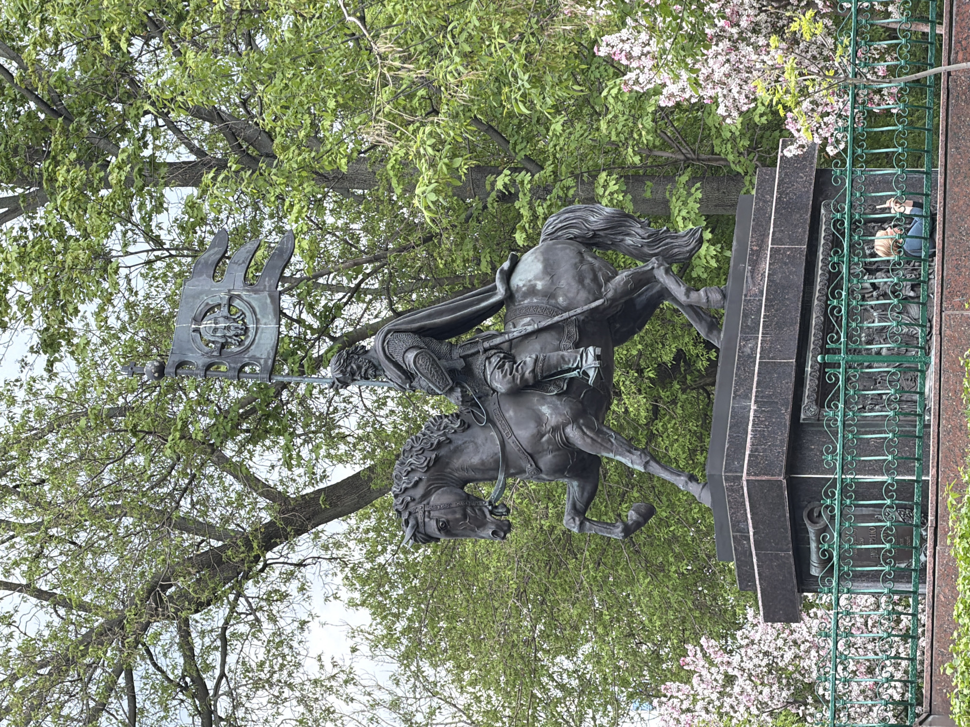 The image captures the monument of Yuri Dolgoruky, the founder of Moscow, Russia. The bronze statue depicts Yuri Dolgoruky riding a horse, holding a banner with trident emblem. The monument stands atop a dark marble pedestal surrounded by a green metal fence. Lush green trees with some flowering branches create a natural backdrop. In the background, behind the monument, two people are visible by the fence.