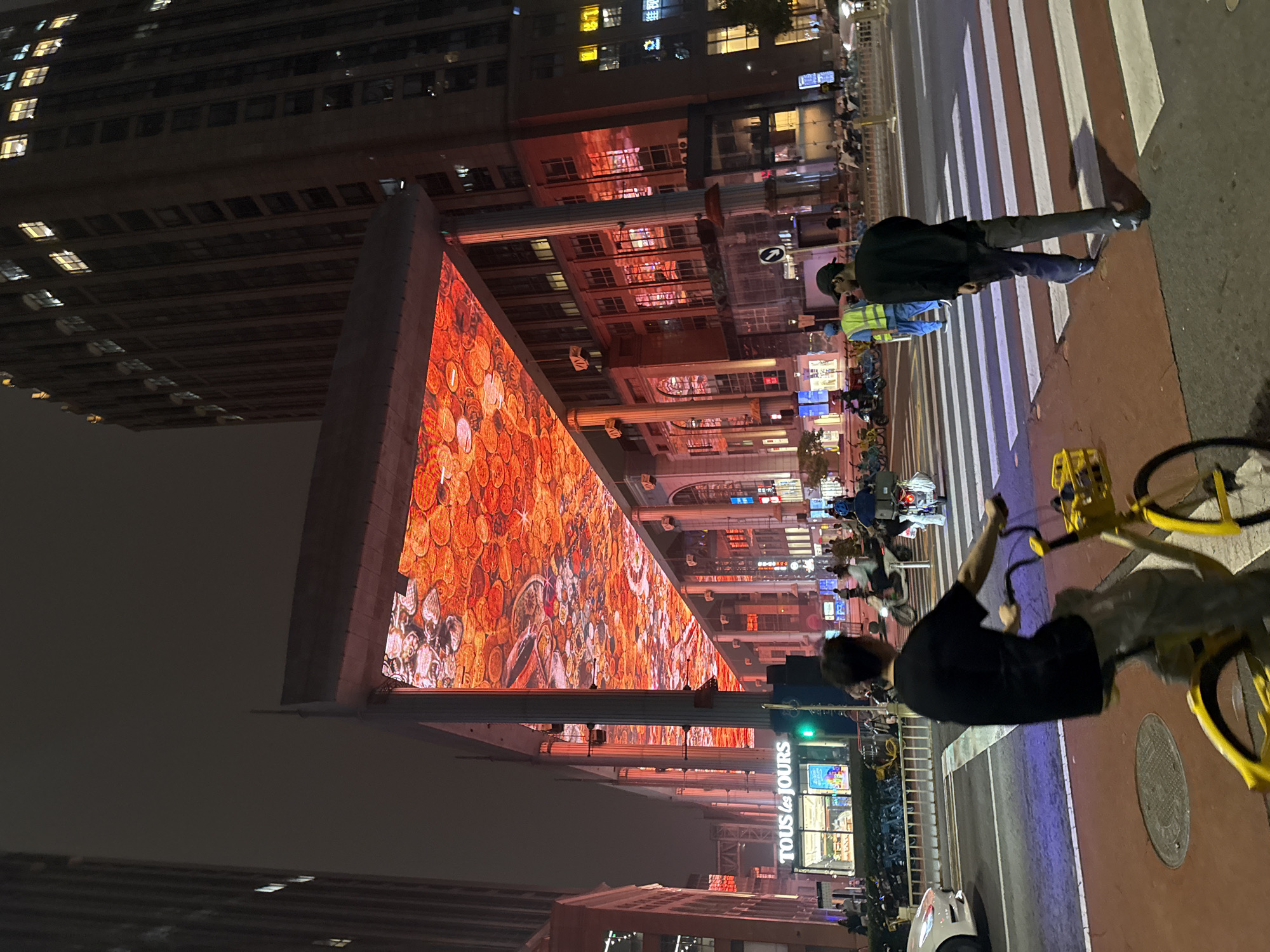 This is a nighttime street scene in Chaoyang District, China. The image features a wide pedestrian crossing with white stripes on a red asphalt surface. A man in a black t-shirt and dark pants is walking across the crosswalk from right to left, his back to the camera. To his left, another person is riding a yellow bicycle with a yellow basket in the foreground, also moving from right to left across the crosswalk.
In the background, a large building with many illuminated windows towers over the scene. Above the street level, a massive LED screen displays a vibrant orange and red pattern, illuminating the underside of a covered walkway. A "TOUS LES JOURS" bakery sign is visible on the left side of the street. Other people, motorcycles, and bicycles are present on the street and sidewalks, with streetlights and building lights contributing to the overall illumination of the urban environment. The sky is dark and overcast.