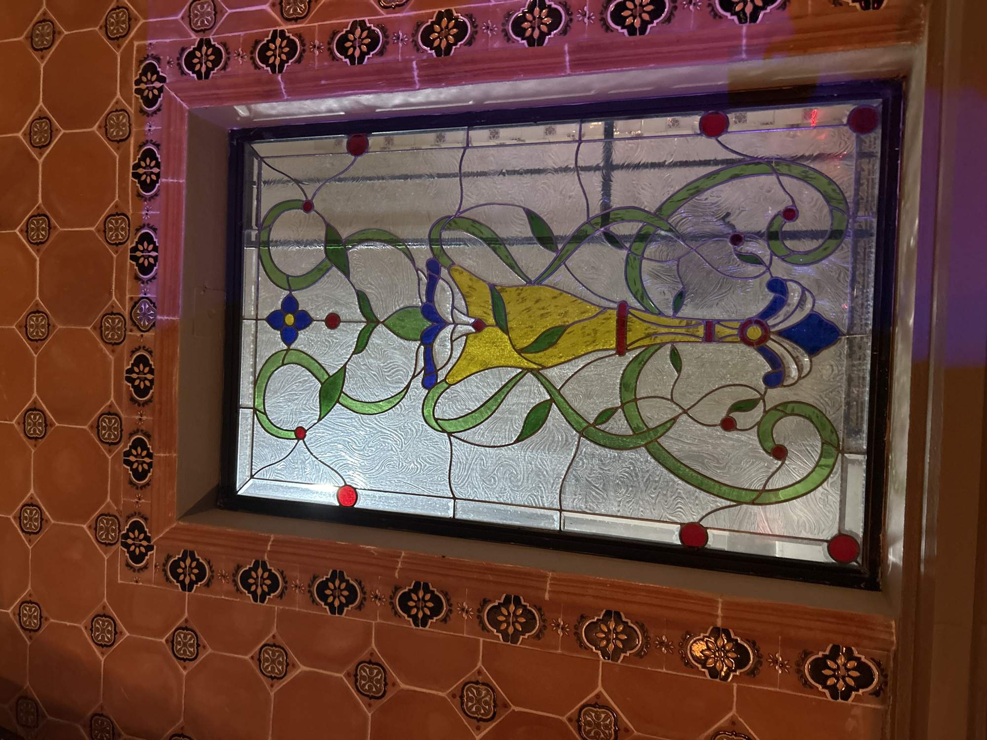 A stained-glass window is surrounded by terracotta-colored square tiles, some decorated with a stylized flower design, and detailed borders with alternating light and dark floral motifs. The window itself features a translucent background with decorative swirling patterns, overlaid with a design incorporating green vines, blue and yellow flower-like elements, and red circular accents. The window frame is black, and the whole assembly is set into a wall. The location is Santa Ana Yancuitlalpan (Chilhuacán), Mexico.
