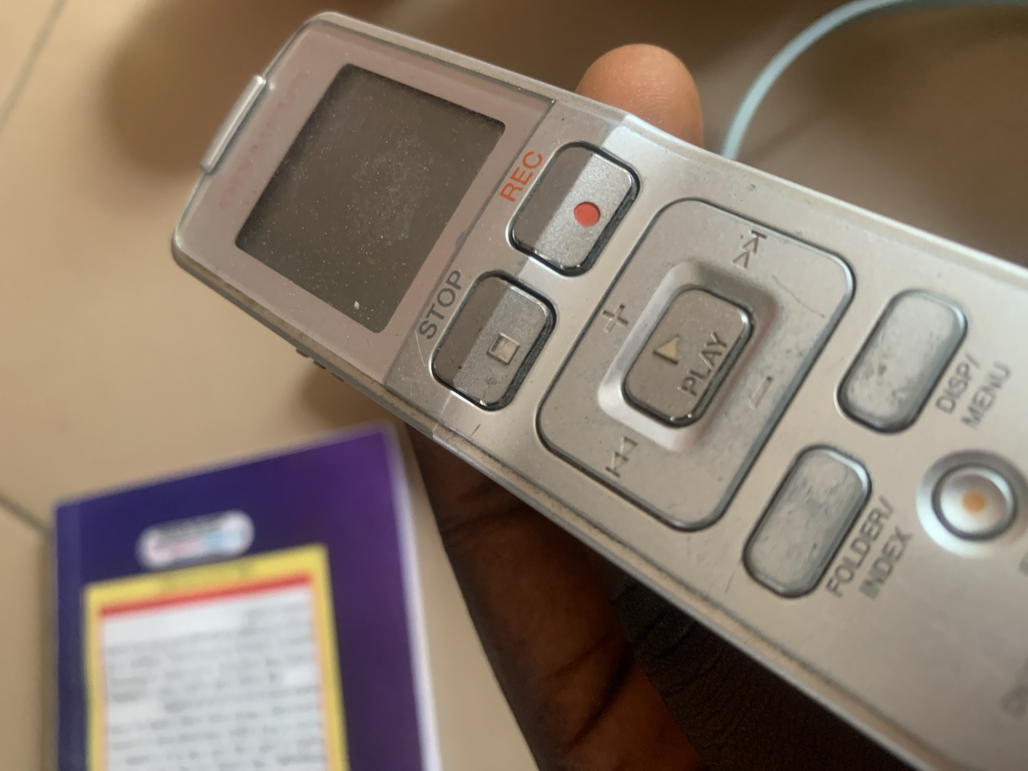 A close-up, eye-level, indoor shot shows a digital voice recorder held by a person's dark hand. The recorder is silver and features several buttons and a small screen. The buttons are labeled with text such as "STOP", "REC", "PLAY", "FOLDER/INDEX", and "DISP/MENU". A red circular button is visible below the "REC" label. In the background, out of focus, is a purple object with a yellow and white label. The lighting is adequate, and the focus is sharp on the voice recorder. The image appears to be taken in Jauro Yinu, Nigeria.