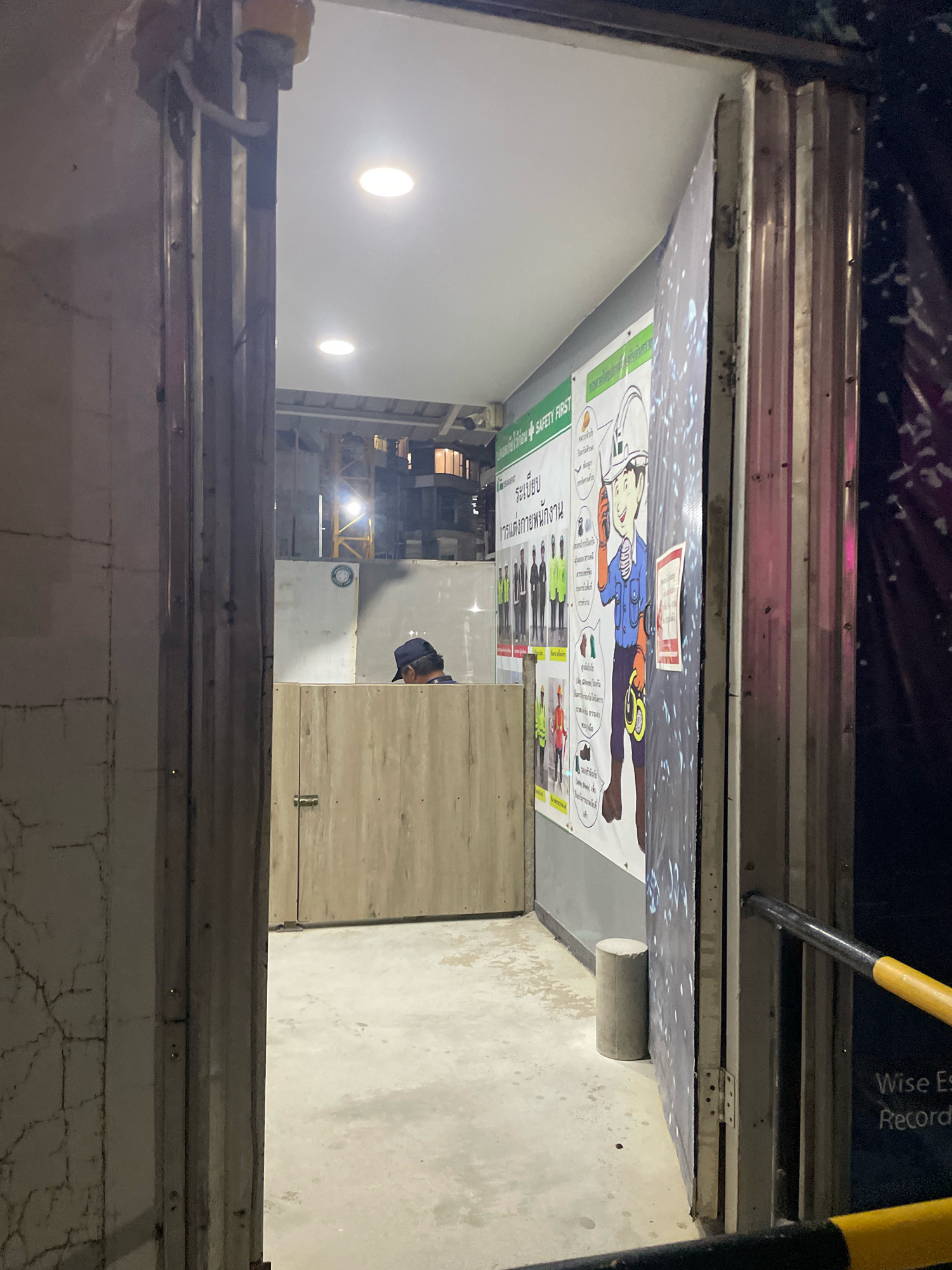 An outdoor view shows the entrance to a small, temporary structure. A man wearing a dark baseball cap is visible inside, seated behind a light-colored wooden counter. To the right of the man, a large safety poster is displayed on a corrugated metal wall, featuring cartoon illustrations of construction workers and safety guidelines in Thai. The poster is partially obscured by a vertical corrugated metal door. The ground is concrete, and a cylindrical concrete bollard stands to the left of the poster. In the background, a construction crane is illuminated, suggesting an ongoing construction project in Pattaya City, Thailand. The scene is dimly lit by interior ceiling lights and the exterior lights from the surrounding area.