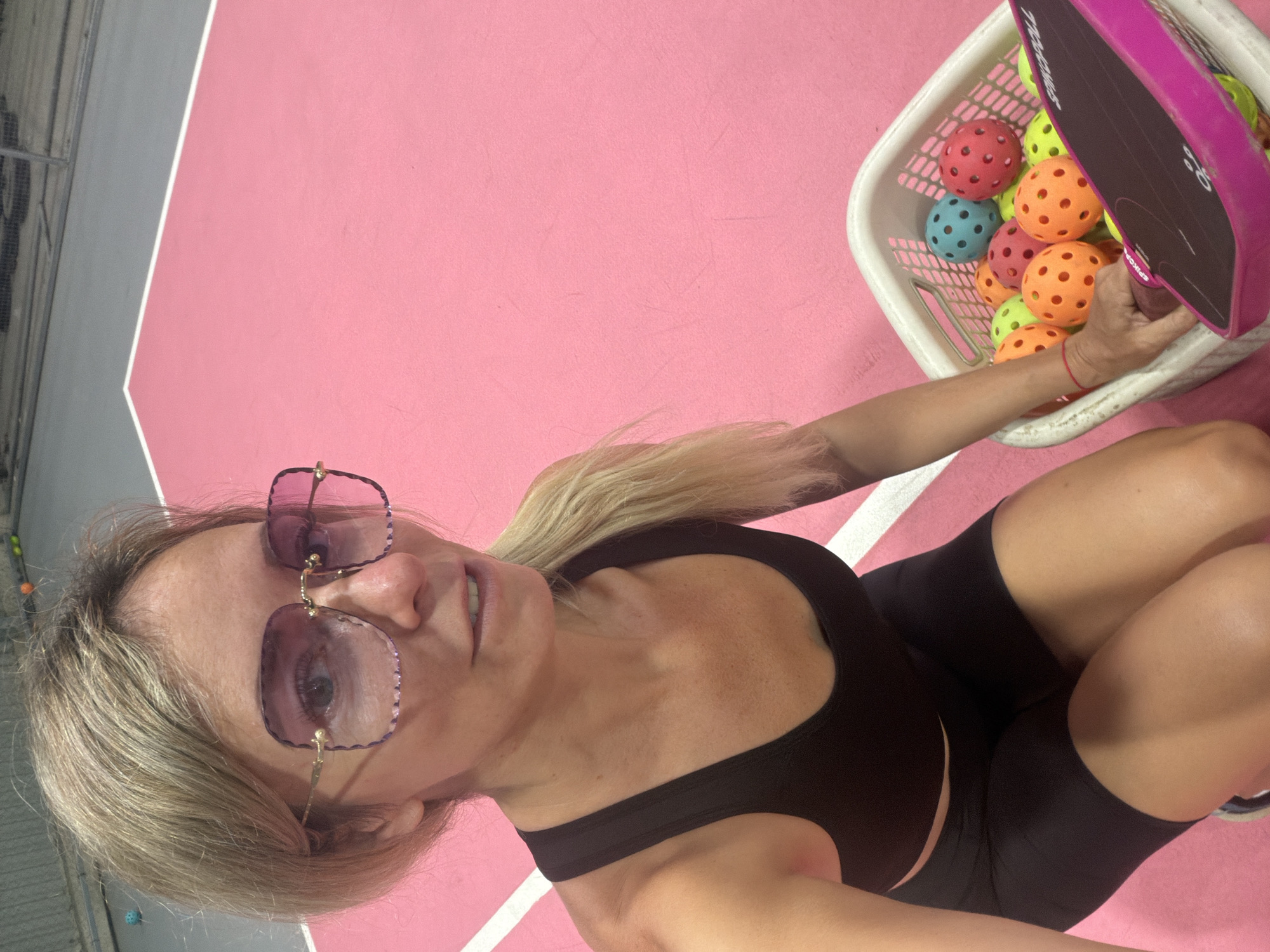A woman with blonde hair and large pink-tinted glasses is kneeling on a pink pickleball court. She is wearing a black sports bra and black shorts. To her right, there is a white basket filled with colorful pickleball balls. She is reaching into the basket to retrieve a paddle, which is pink and black. In the background, parts of the court fencing and other pickleball courts are visible. The scene appears to be an indoor pickleball facility. The location in Da Lat, Vietnam is not provided in the image.