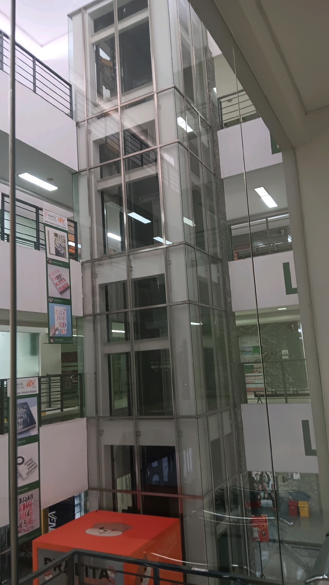The image shows a modern glass elevator shaft in a multi-story building, likely a commercial or public space, in Tangerang, Indonesia. The elevator shaft is a central feature, made of glass panels and metal framing, revealing multiple floors and internal structures. Fluorescent lighting is visible on several floors. On the left side of the shaft, several vertical banners with text and graphics are hanging, suggesting promotional or informational displays. In the foreground at the bottom, a large orange cube with a cartoon-like image of an animal face is visible. Other architectural elements such as railings, walls, and windows are present, indicating the interior of a building. The overall impression is of a contemporary indoor environment with a focus on vertical circulation.