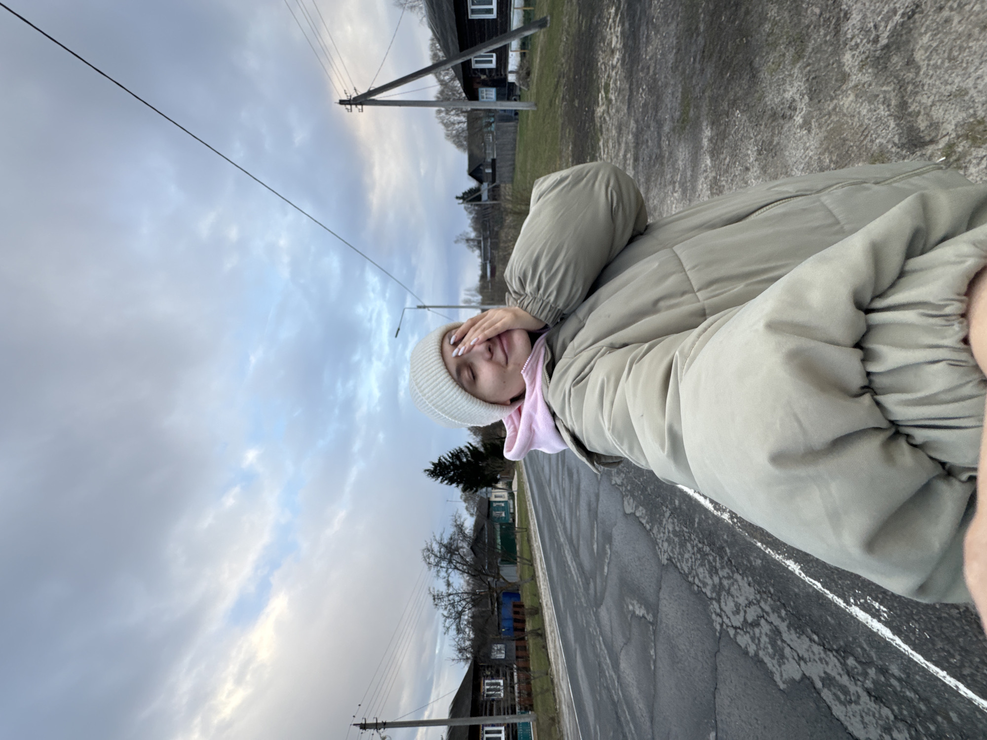A person is standing outside along a road in Красный Маяк, Russia. They are wearing a light-colored beanie, a pink hoodie, and a light-colored puffer jacket. The person has one hand covering their face. Behind the person is a road with cracks, followed by a row of houses. Utility poles and wires are in the background. The sky is overcast.