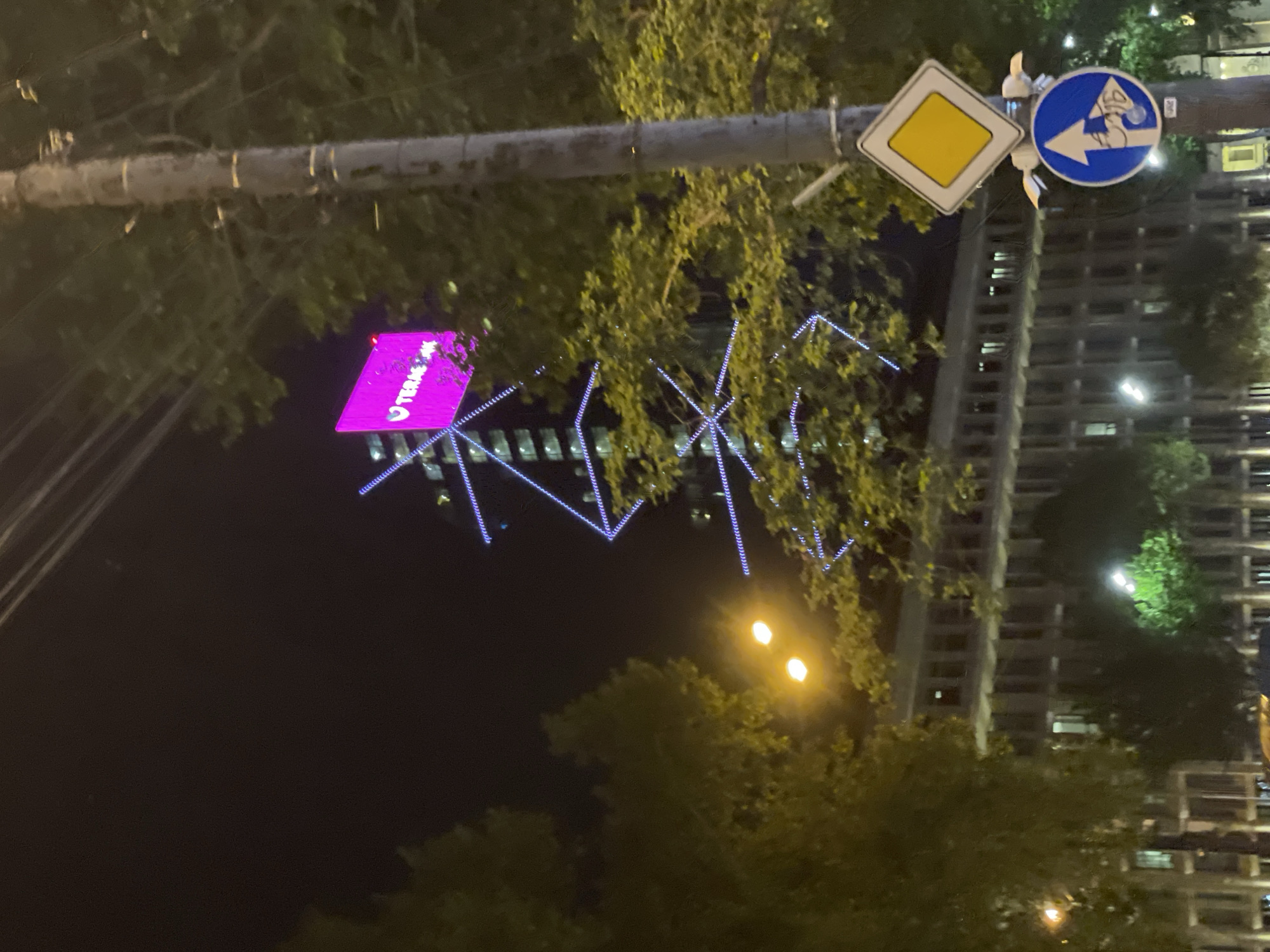 The image shows a night scene in Tbilisi, Georgia. In the foreground, there are street signs for road conditions and direction. The sky is dark. Behind the signs is an illuminated building decoration. Streetlights illuminate the scene. Trees obscure portions of the building in the background. A pink sign with white text is visible. Blue lights form a decorative pattern on the building.