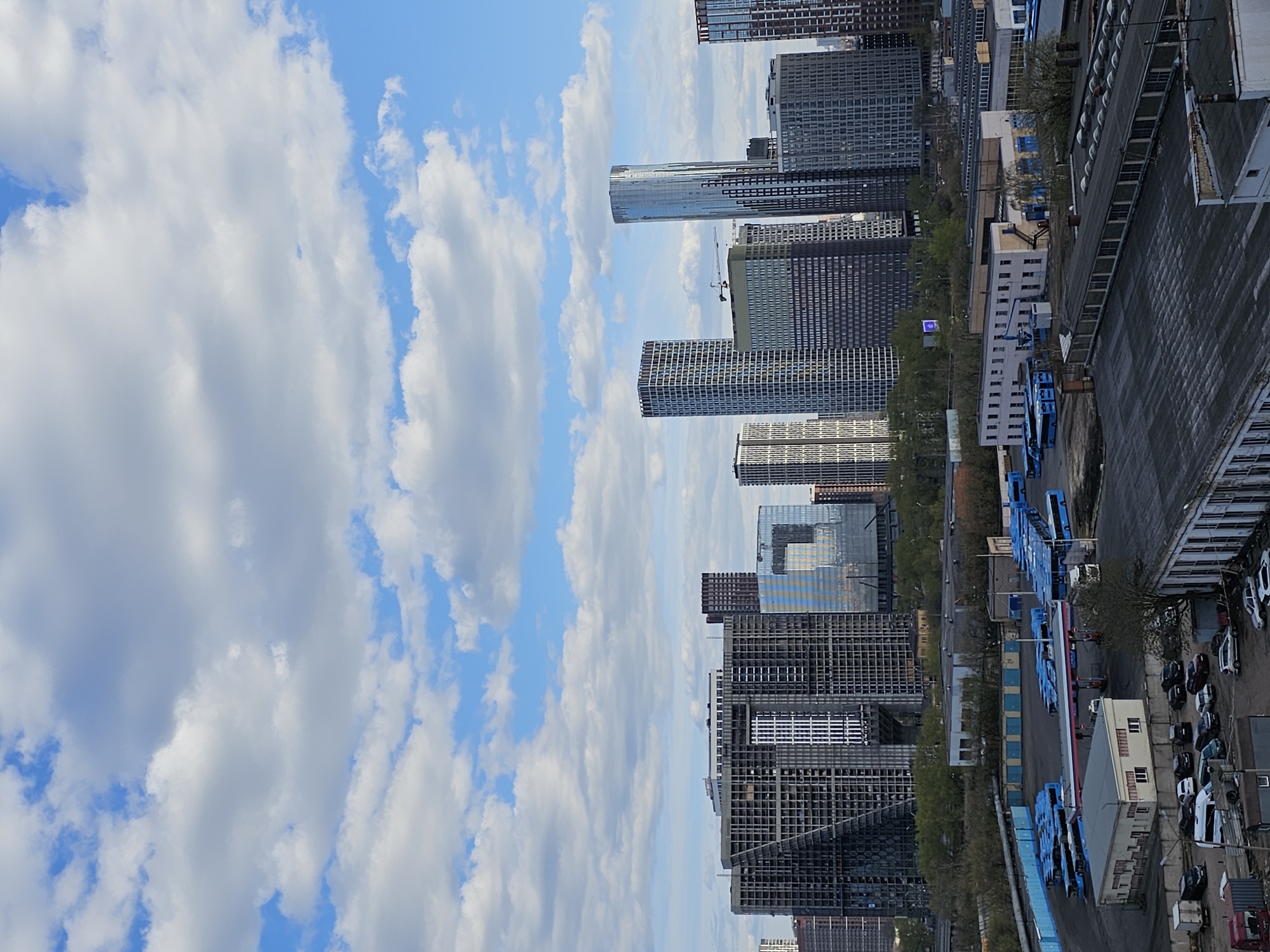 The image presents a panoramic view of an urban landscape in Moscow, characterized by a stark contrast between modern high-rise architecture and more functional, industrial-style buildings. The scene is set under a bright, partly cloudy sky, with patches of clear blue visible between large, fluffy white cumulus clouds, suggesting a clear daytime.
In the mid-ground and background, a cluster of numerous contemporary skyscrapers dominates the skyline. These tall buildings exhibit sleek designs, primarily constructed with glass and metal facades, some featuring distinct grid patterns. One prominent slender, cylindrical high-rise stands out, and a tall construction crane is visible amidst these towering structures, indicating ongoing urban development.
The foreground and closer mid-ground reveal a more industrial or commercial zone. A large paved area serves as a parking lot, filled with dozens of various cars and several long rows of distinctive blue vehicles, possibly buses or specialized utility trucks. Adjacent to these vehicles, a gas station with a red and white striped canopy is visible. Scattered throughout this area are several lower-rise commercial or industrial buildings, including a large, dark-roofed structure with multiple vents, likely a warehouse or factory. Some smaller white buildings with multiple windows are also present, interspersed with pockets of green foliage and trees.
The scene appears static, with no discernible people or active movement of vehicles. No visible text or specific emotional cues are present. The overall impression is one of a dynamic city undergoing modernization while maintaining its commercial and industrial operational aspects.