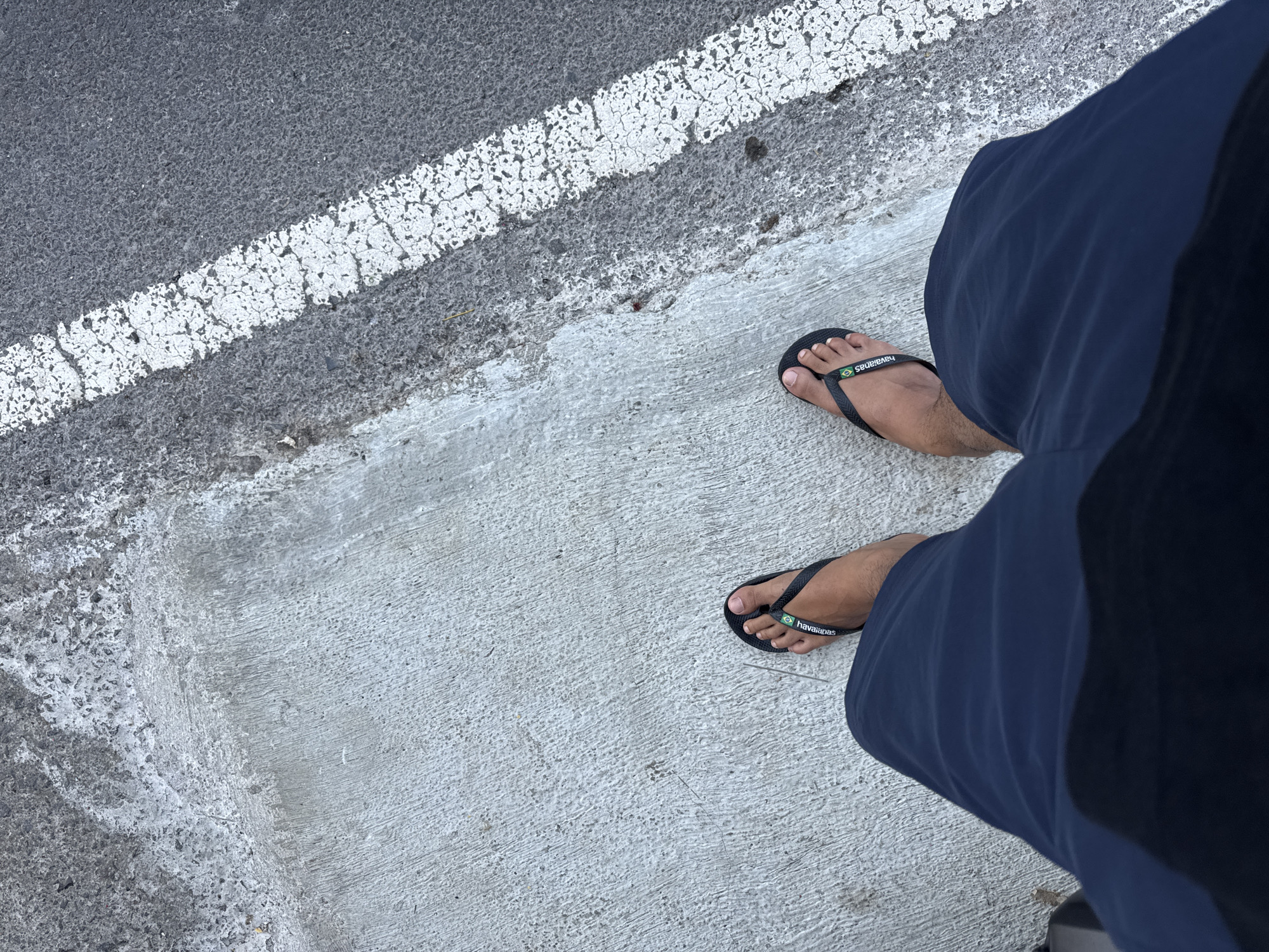 An overhead view shows a person standing on a sidewalk next to a road. The person is wearing dark blue shorts and black flip-flops with the brand name "Havaianas" visible on the strap of the left flip-flop. The sidewalk is made of concrete with a rough texture. The road adjacent to the sidewalk is asphalt and marked with a white line. The location is Busungbiu, Indonesia.